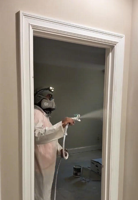 A worker wearing protective gear, including a helmet and mask, sprays disinfectant or paint on a doorway frame inside a room.