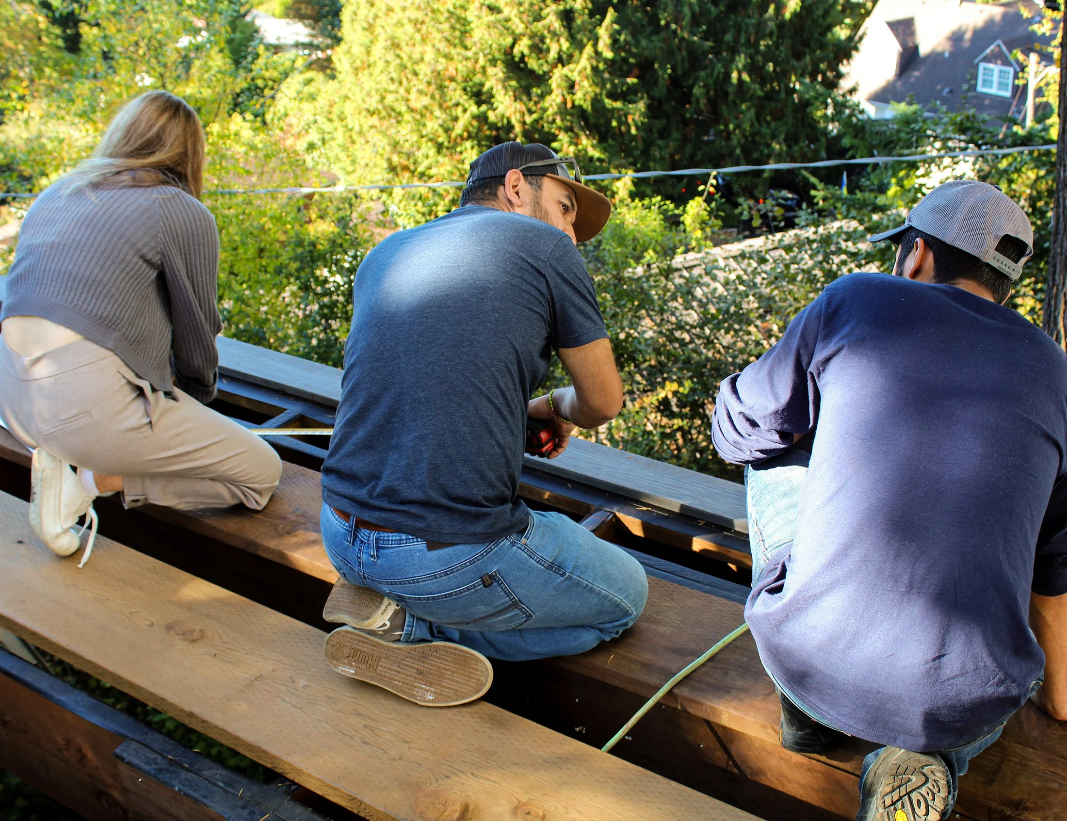 Three people, two men and one woman, are seated outdoors on a wooden table, with green trees and houses in the background. The woman is on the left side, wearing a gray sweater and beige pants. The man in the middle wears a dark blue shirt, glasses, and a baseball cap. The young man on the right is wearing a gray cap and a dark blue shirt.