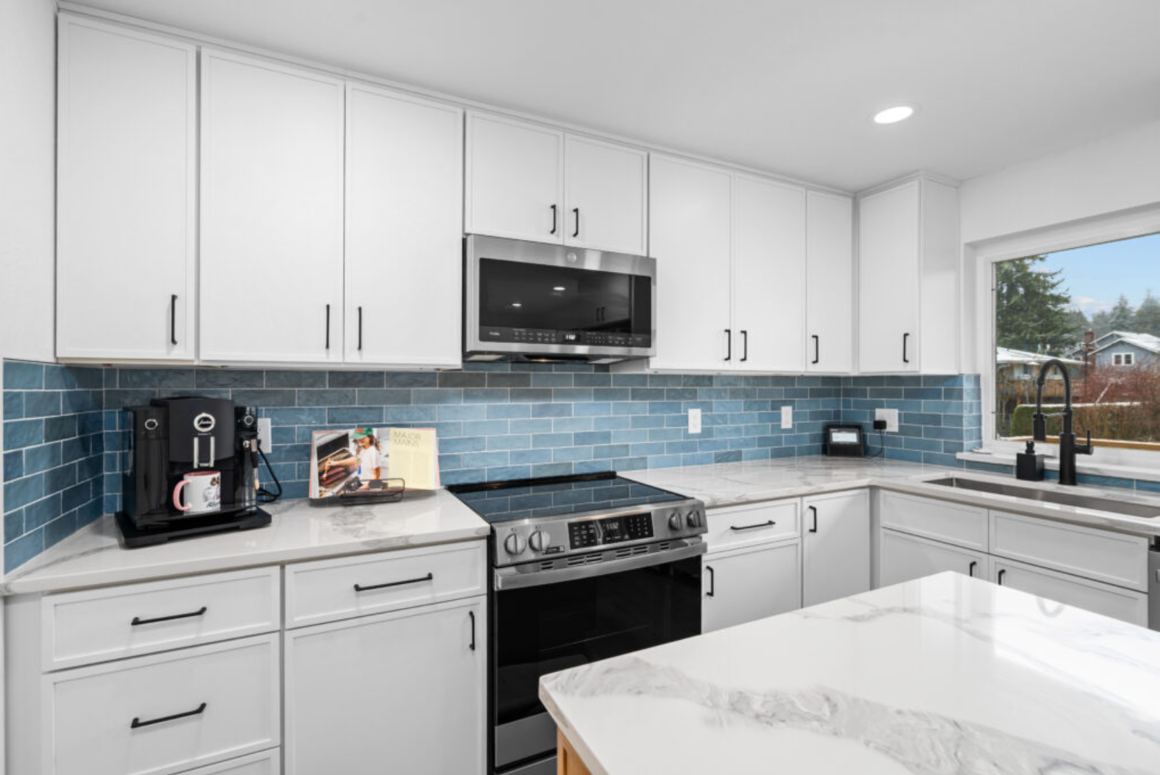 Kitchen with white cabinets, black handles, blue subway tile backsplash, white marble countertops, stainless steel microwave and oven, coffee maker, window above sink, black faucet, and outdoor views.