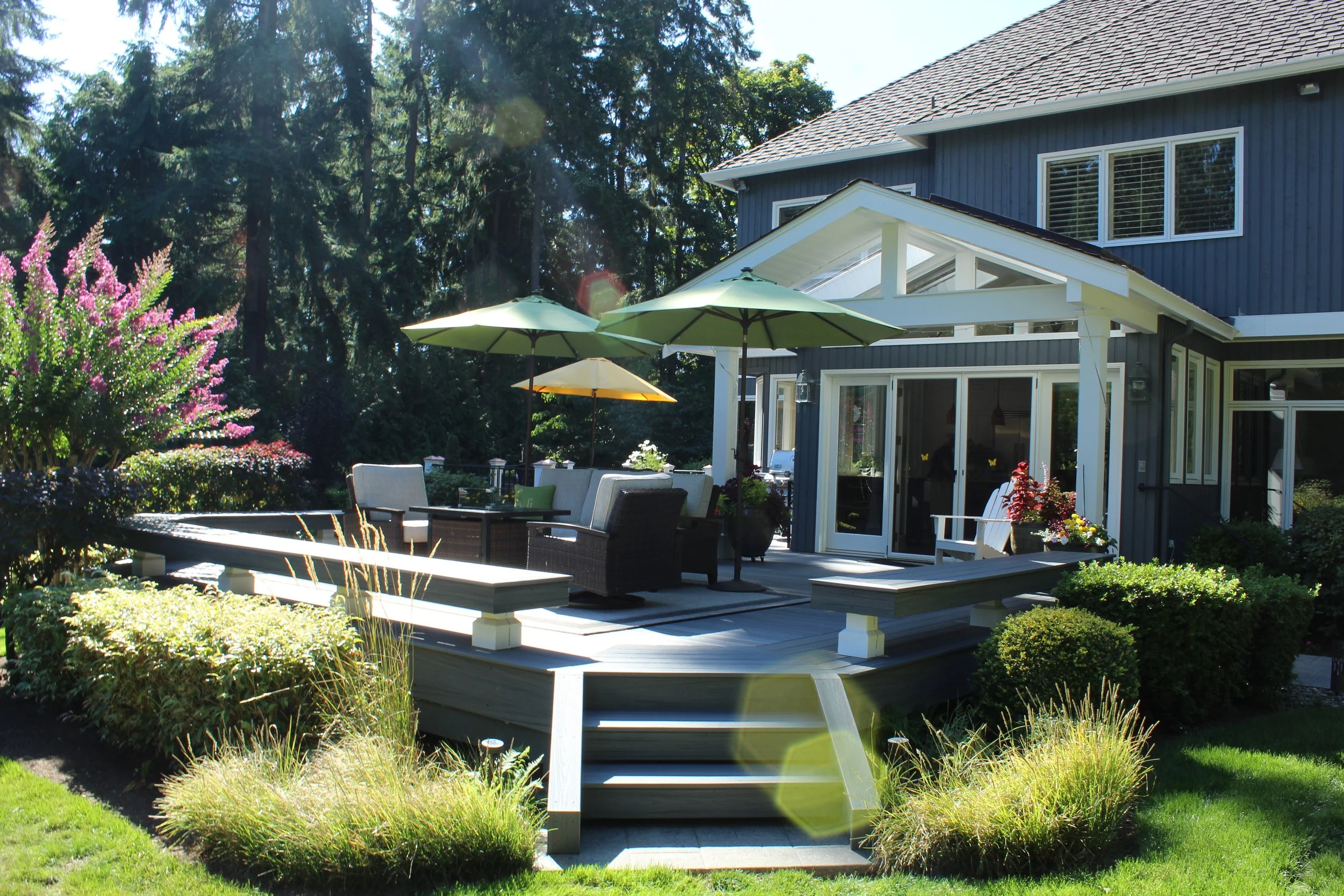 A backyard patio with outdoor furniture, umbrellas, and surrounding greenery, adjacent to a blue house with large windows, on a sunny day.