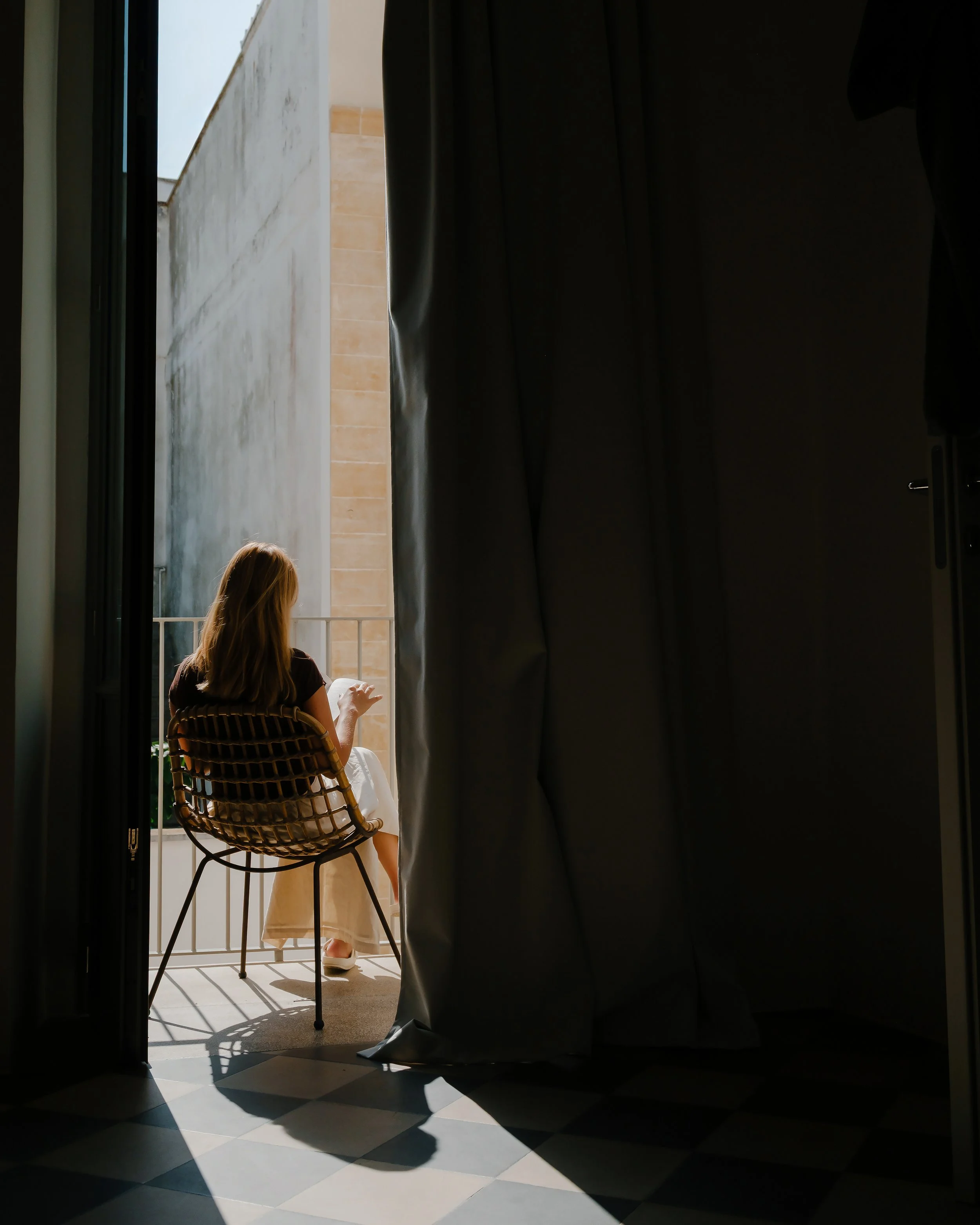 A woman sitting on a patio chair, holding a cup, is visible through an open door, with sunlight illuminating her and casting shadows on a tiled floor.