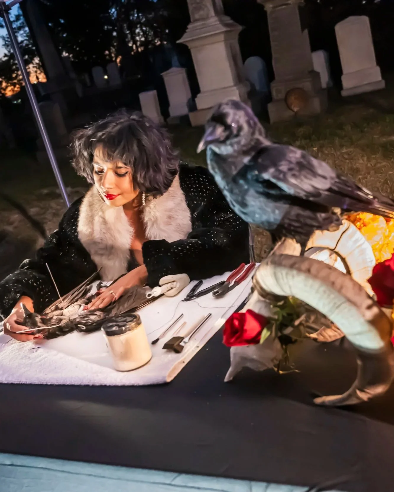 A woman with wavy gray hair and red lipstick is sitting at a table decorated for a celebration or event, surrounded by flowers, feathers, and a small black and white kitten. She is looking down at the kitten, and a large bird with gray and black feathers, possibly a magpie, is perched close to her on the table. The background features gravestones, indicating they are in a cemetery during dusk or early evening.