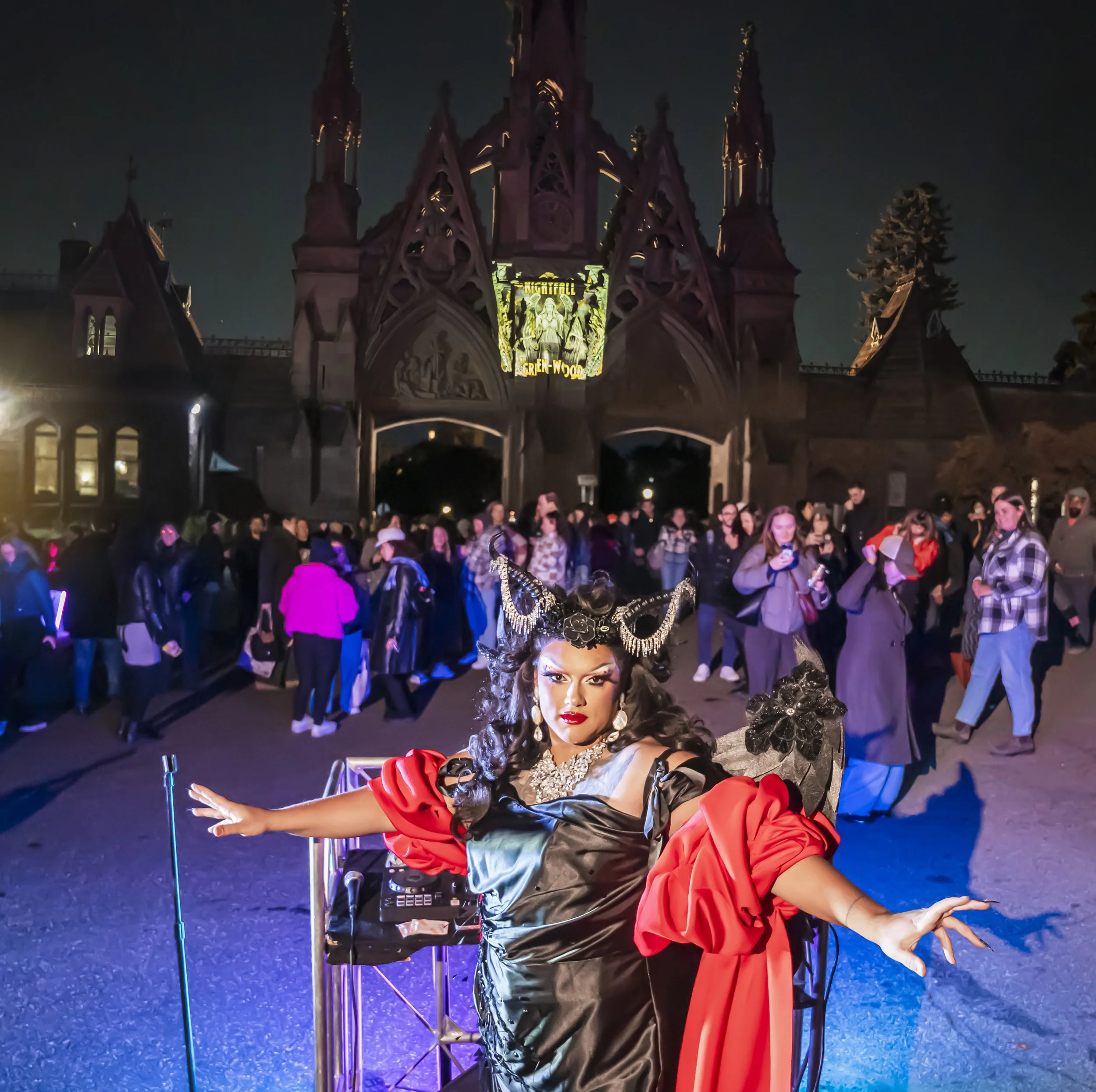 A performer in vibrant costume poses in front of a castle themed backdrop at night with a crowd watching.