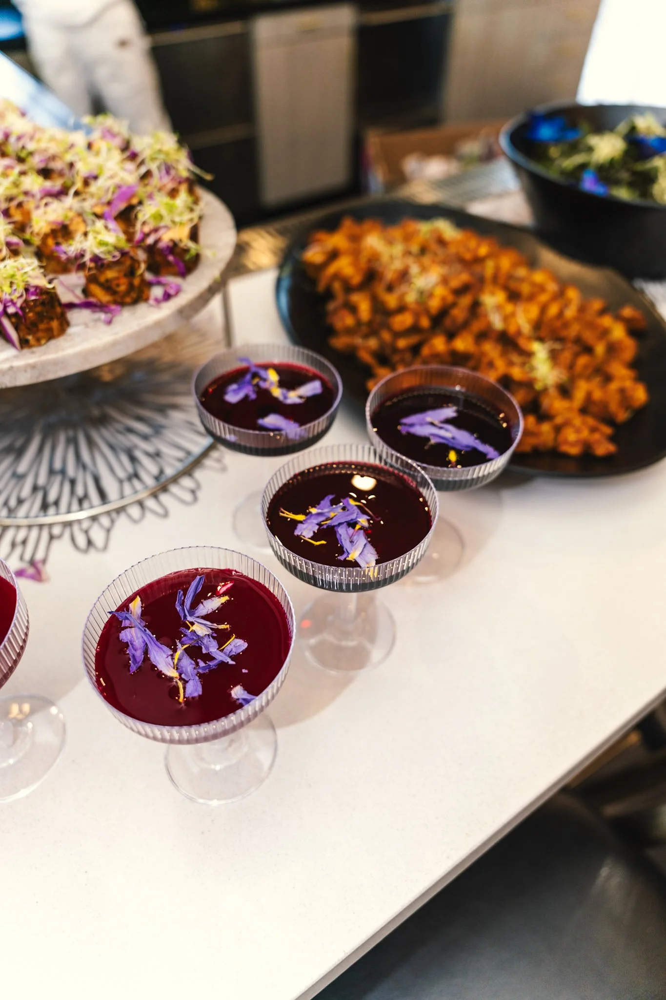 Multiple glasses of non alcoholic elixirs garnished with purple flowers on a white table, with dishes of salad and plant-based cuisine in the background.