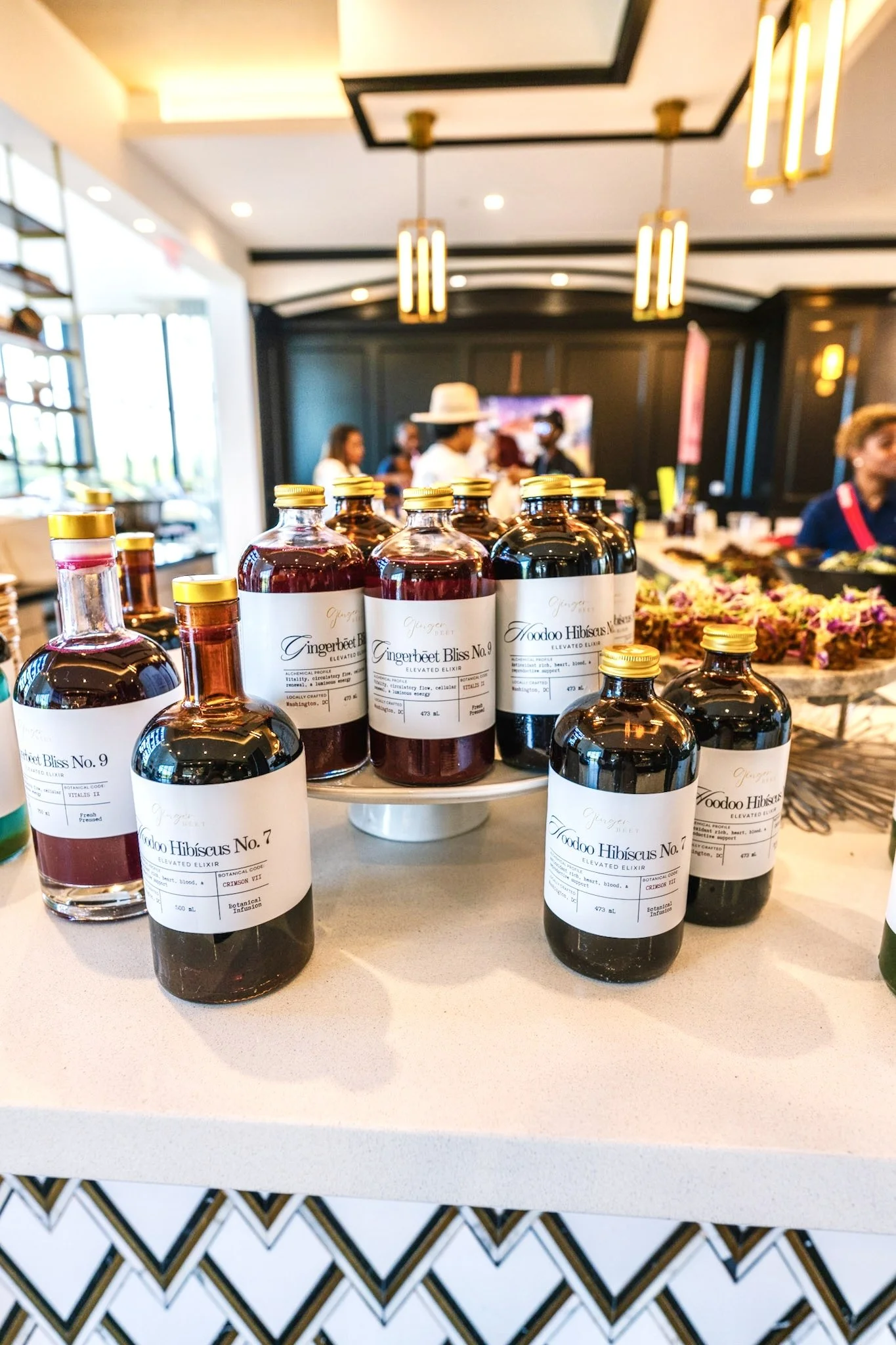 Bottles of tonic labeled Gingerbeet Bliss No. 9 & Hoodoo Hibiscus No. 7 on a table at an indoor event with people in the background. Zero proof elevated elixirs from the Ritūal Réserve collection at Aston Martin. Luxury wellness, plant-based cuisine