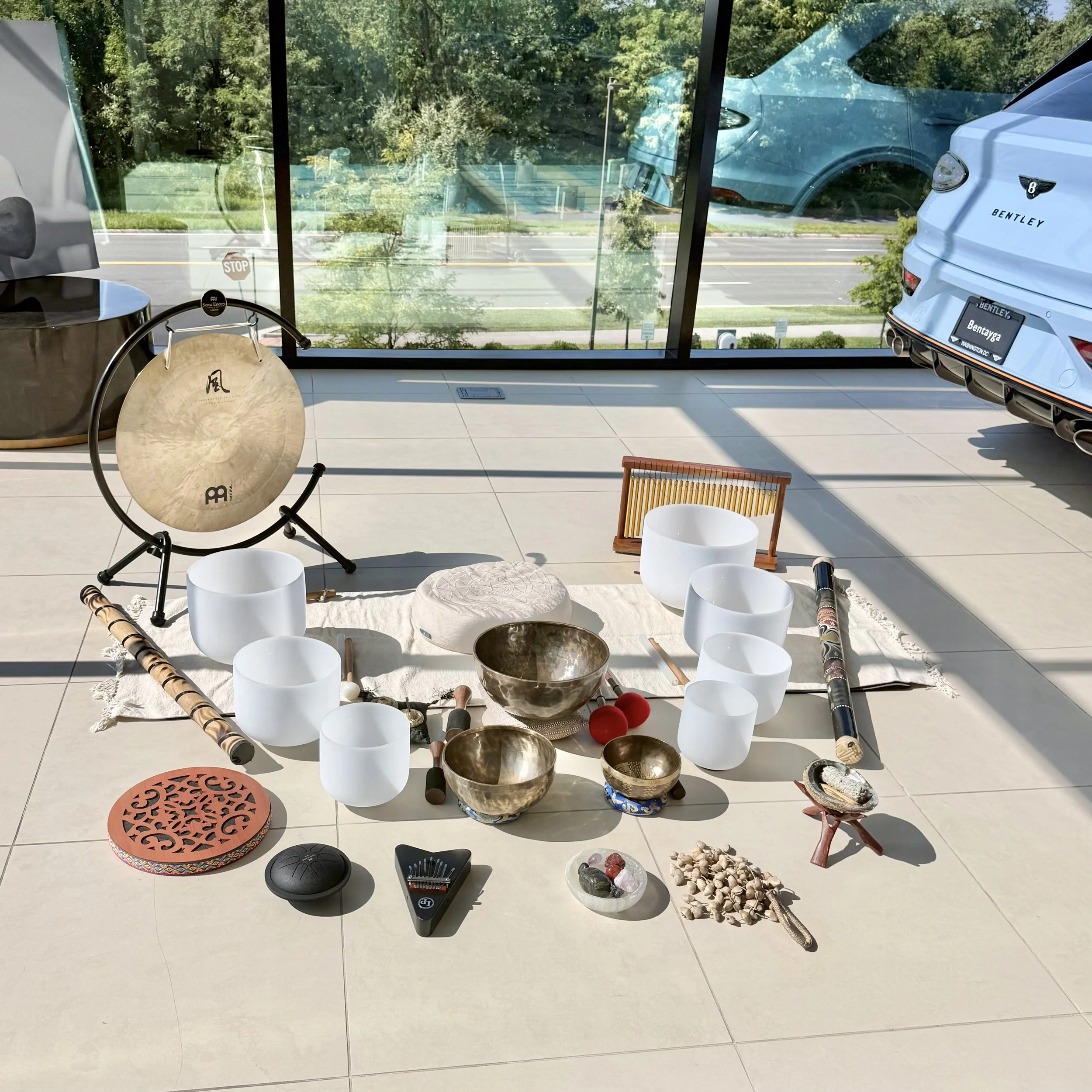 A display of musical instruments and objects on the floor near a large window. Included are a cymbal, a drum, an ocarina, maracas, rocks, a small drum at Aston Martin Bentley showroom floor Washington, DC. Luxury Wellness