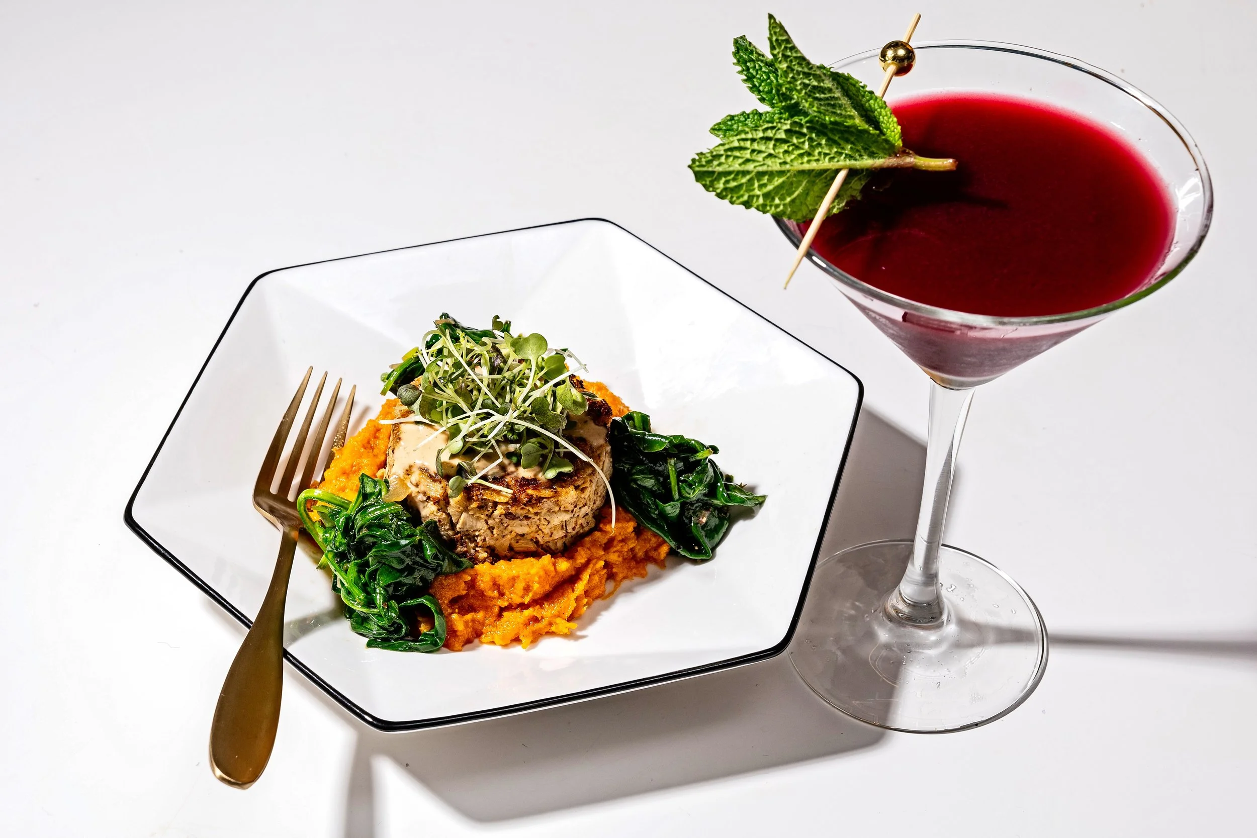 A restaurant plate with a vegan crab cake topped with microgreens, served over mashed sweet potatoes and sautéed spinach, alongside a tall stemmed glass of zero proof mocktail garnished with a mint sprig and a cocktail pick.