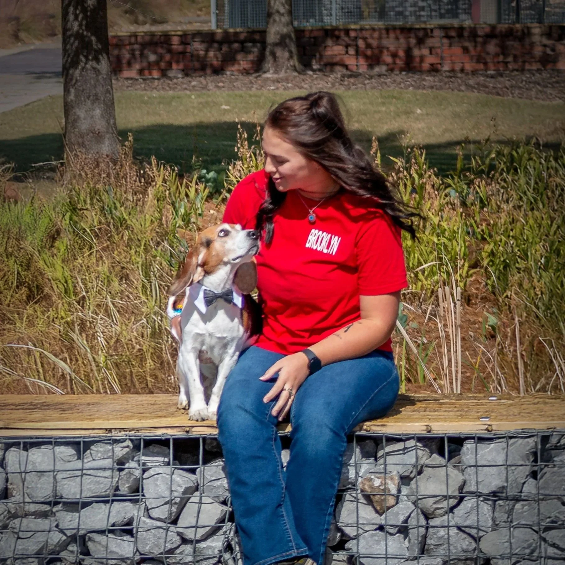 A woman in a red Brooklyn t-shirt is sitting on a bench outdoors, looking at a beagle puppy wearing a tuxedo outfit, in front of a wire fence and a brick wall.