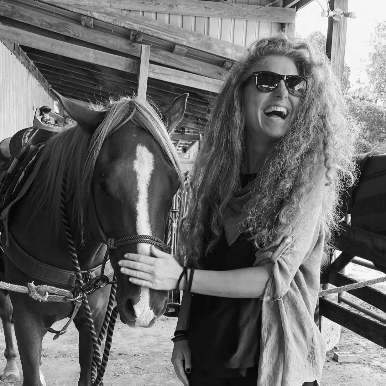 A woman with long, curly hair and sunglasses smiling and touching a horse's face inside a barn or stable.