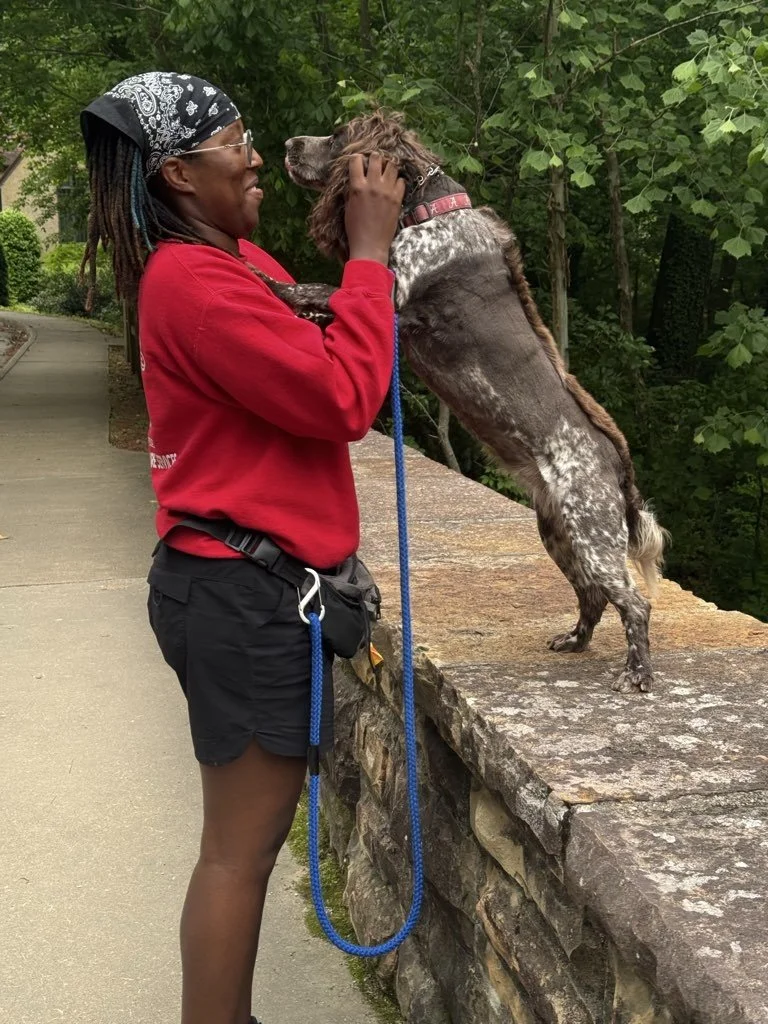 Woman holding and kissing a dog that is standing on a stone ledge, outdoors with green trees in the background.