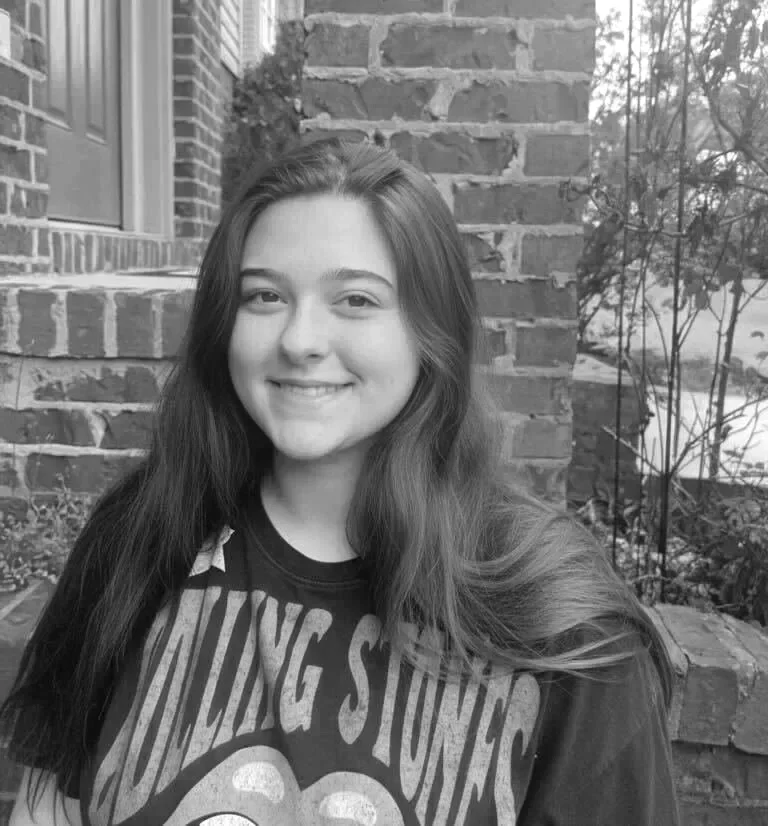 A young woman with long hair smiling, standing outdoors in front of a brick wall and plants, wearing a graphic t-shirt.