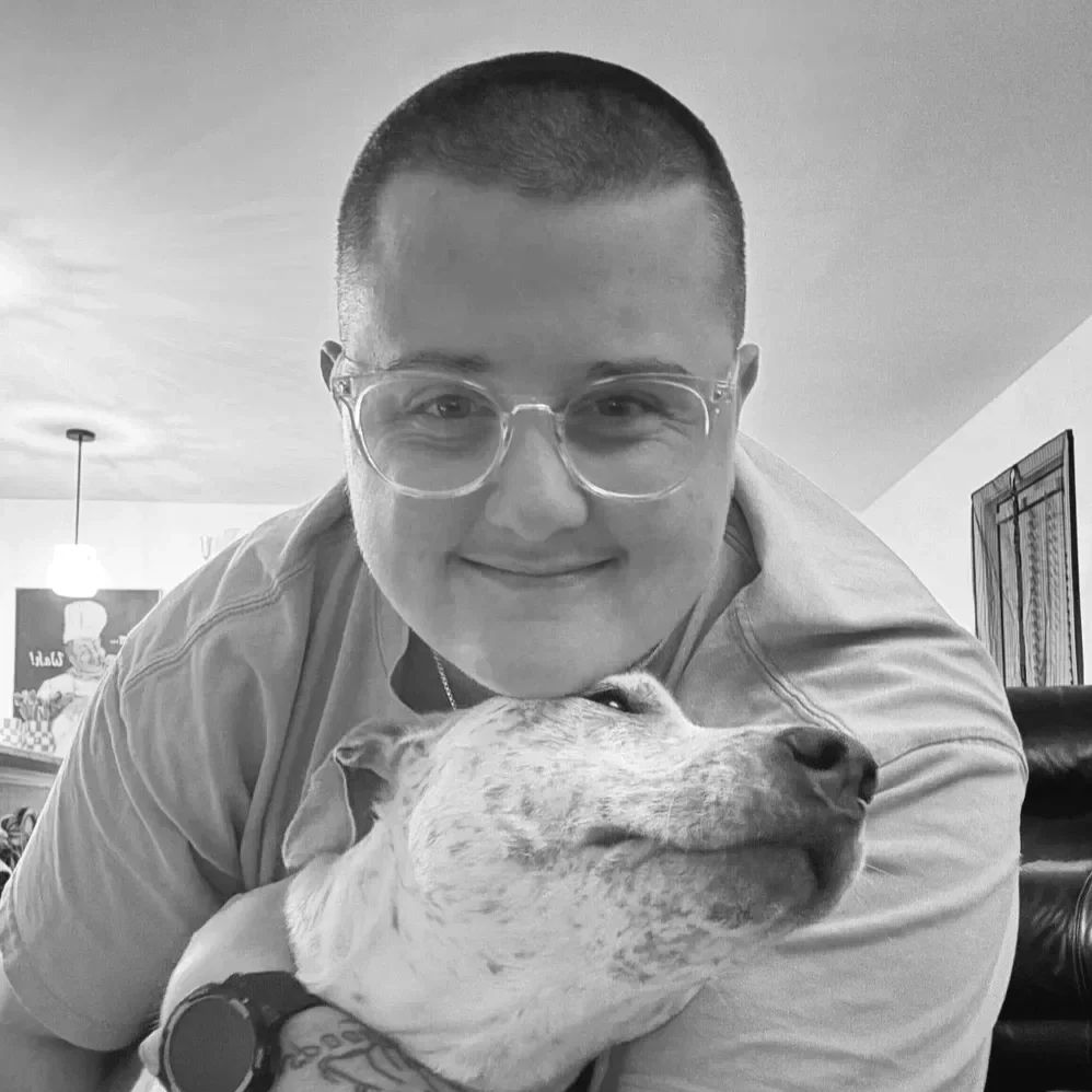 A young person with glasses and a short haircut hugging a dog with a content expression indoors.