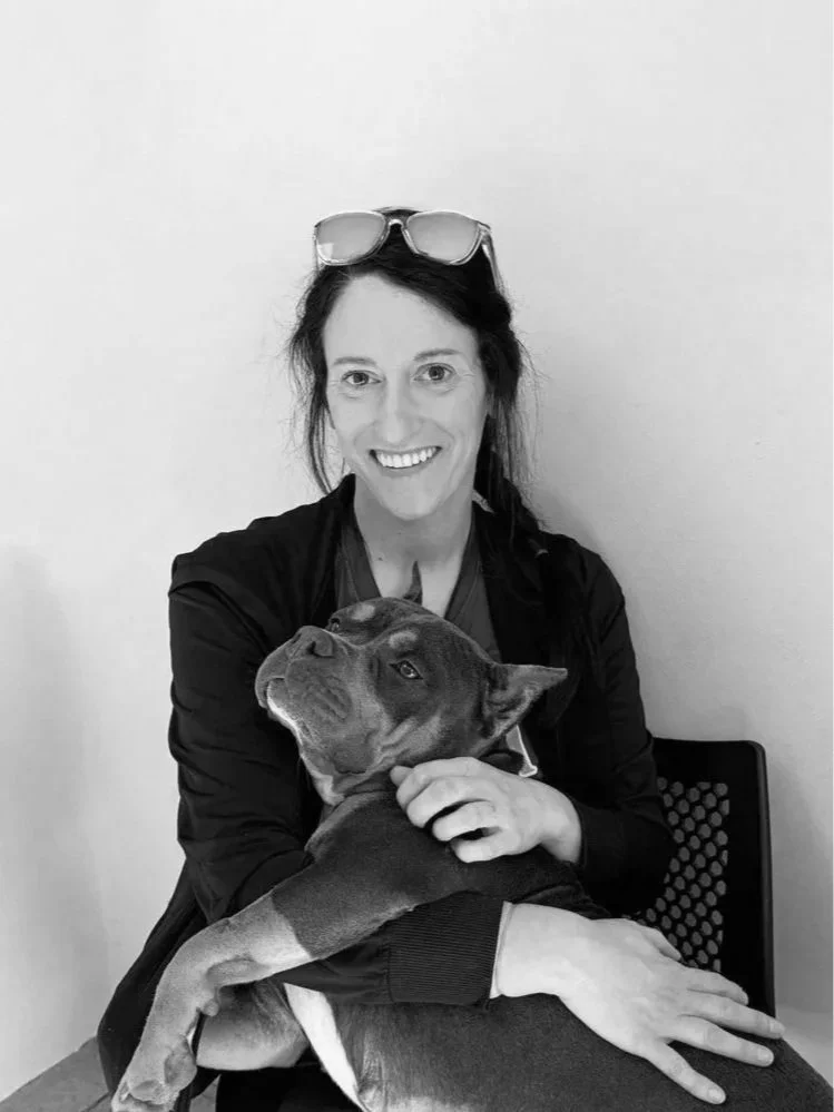 Black and white image of a woman, Kalea, sitting with a dog laying with her.
