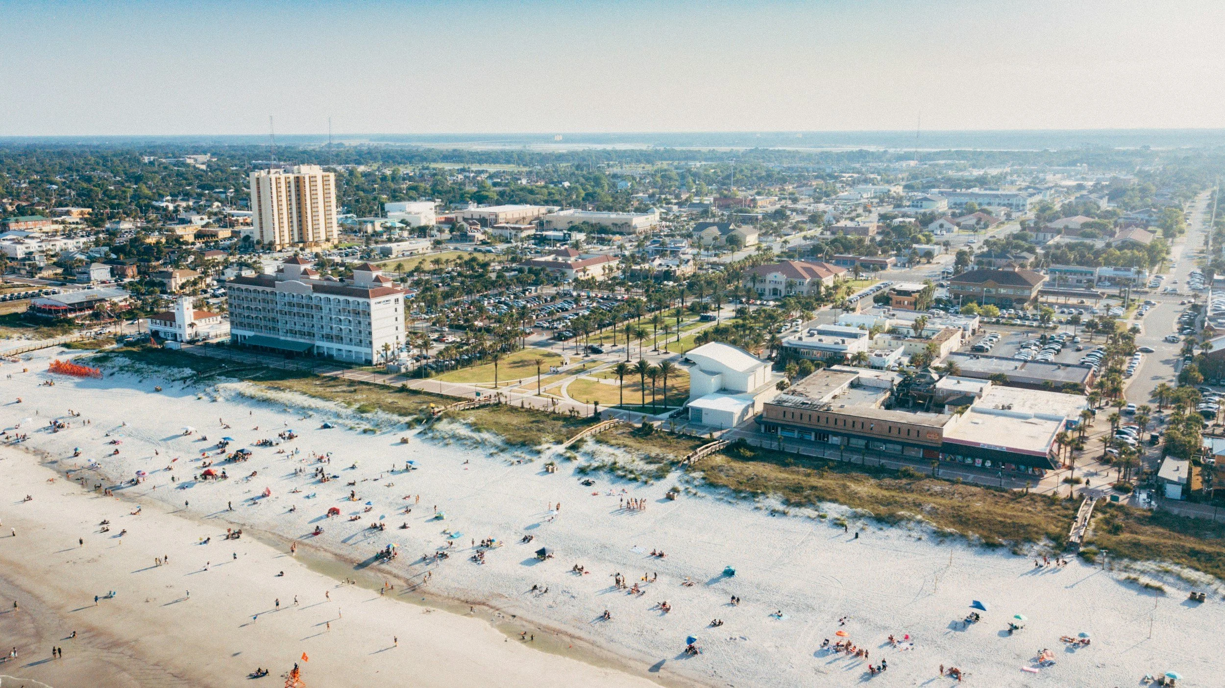 Aerial view of a beach with sand, umbrellas, and people, adjacent to a city with buildings, parking lots, and streets.