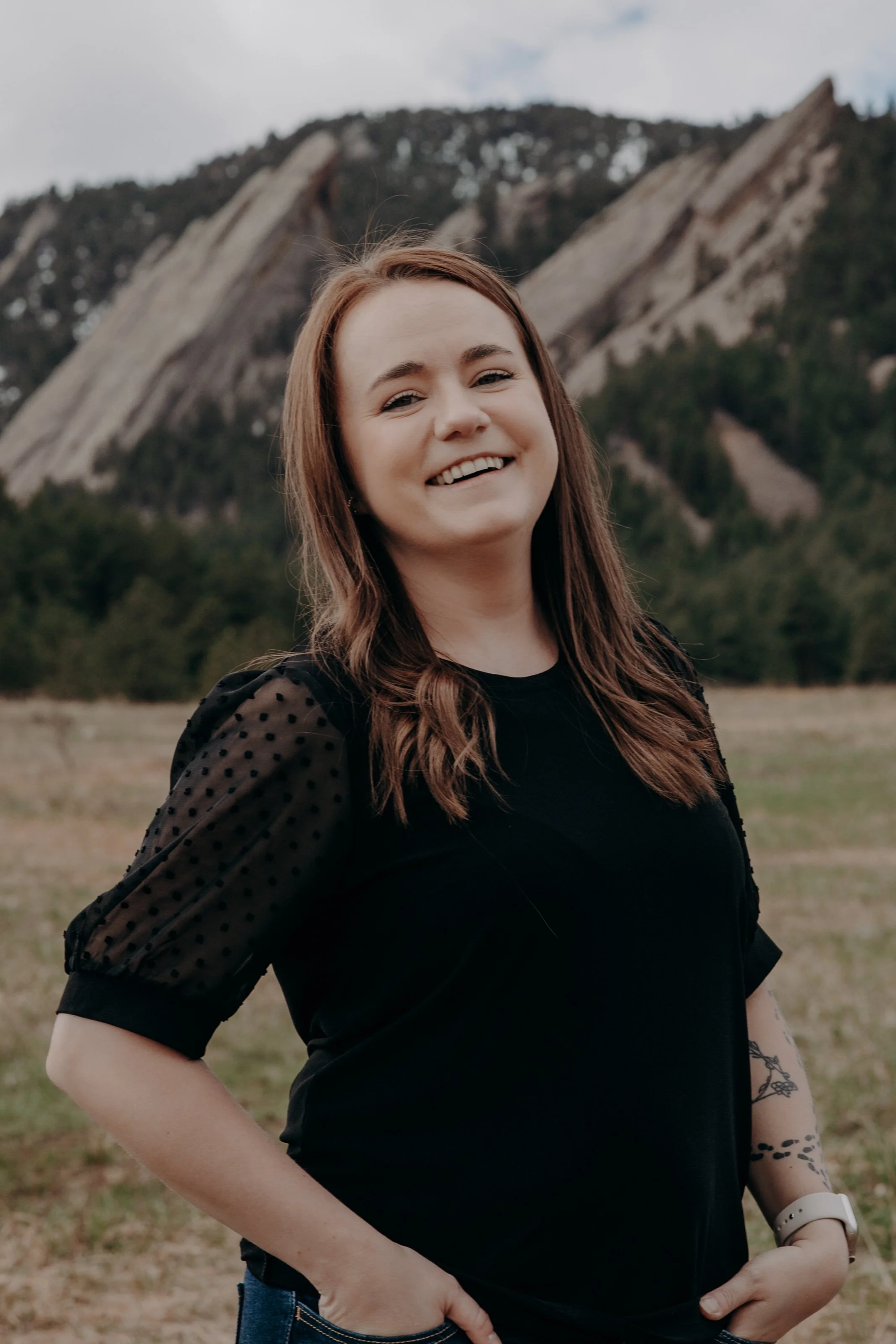 A young woman with long brown hair smiling outdoors, wearing a black top with sheer polka dot sleeves, in front of a mountainous landscape with trees and clouds.