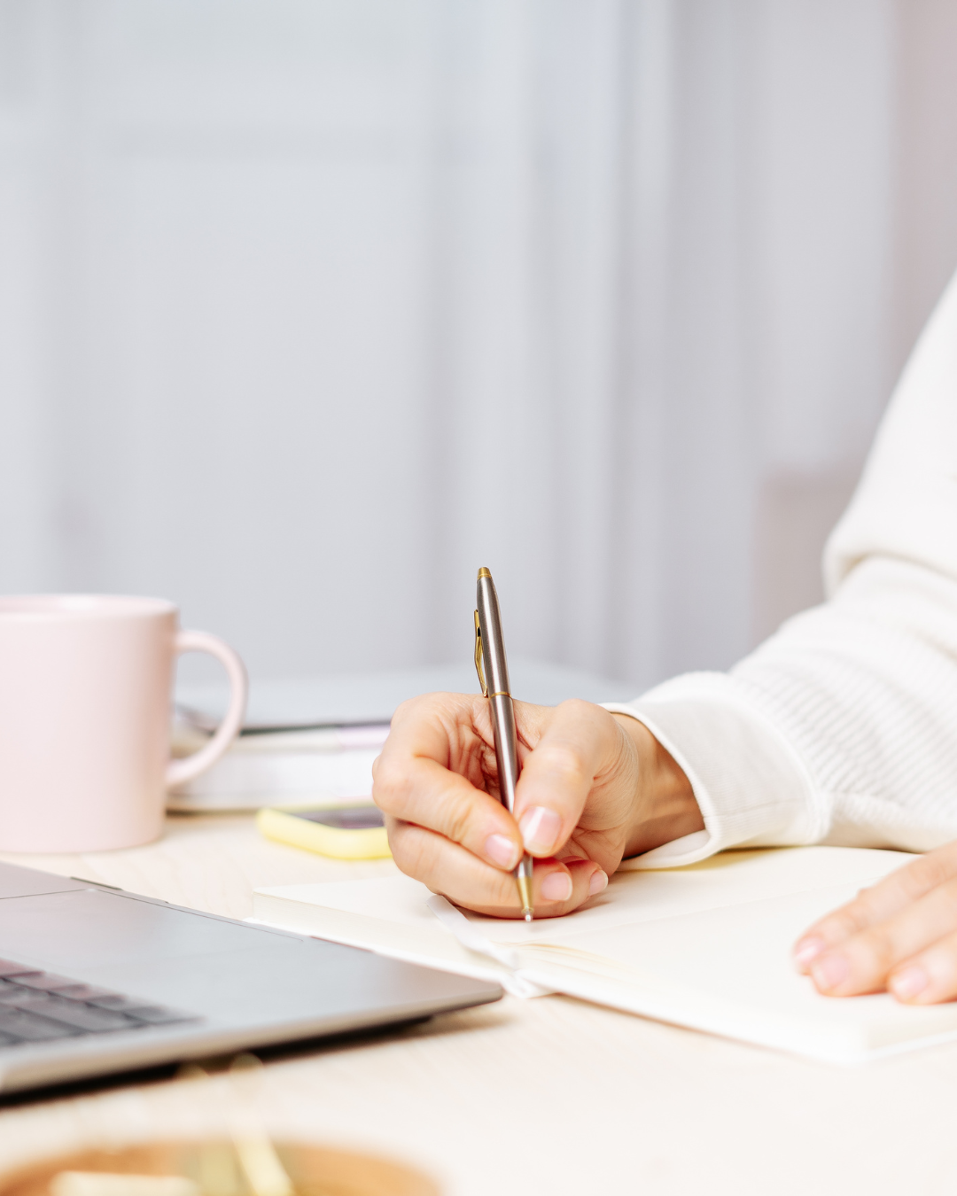 Person writing in a notebook with a pen, a pink mug, and a laptop on a desk.