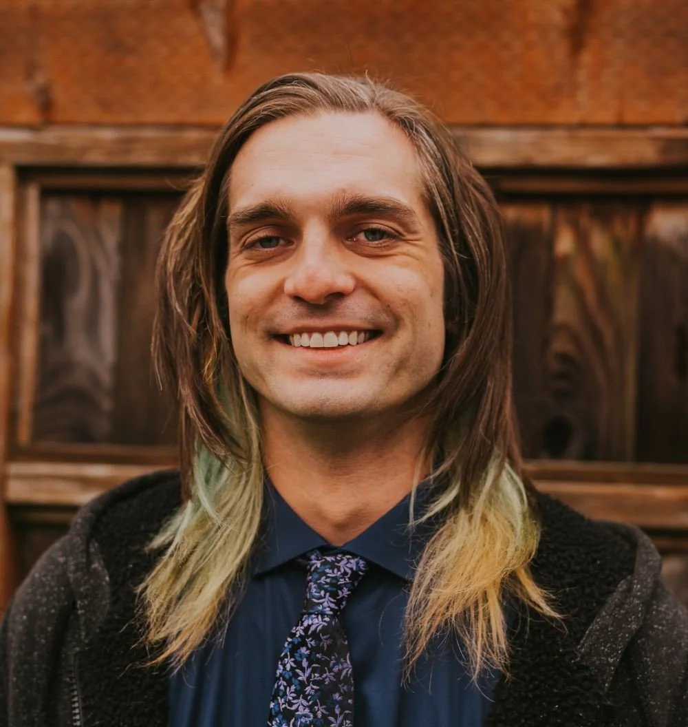 A smiling man with long, wavy hair, wearing a professional navy shirt, and floral tie, standing in front of a wooden background.