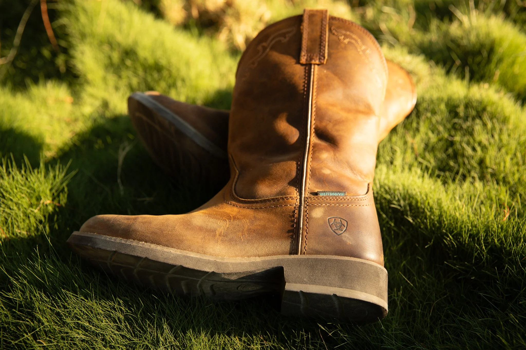 A pair of brown leather waterproof Ariat boots on green grass with sunlight.