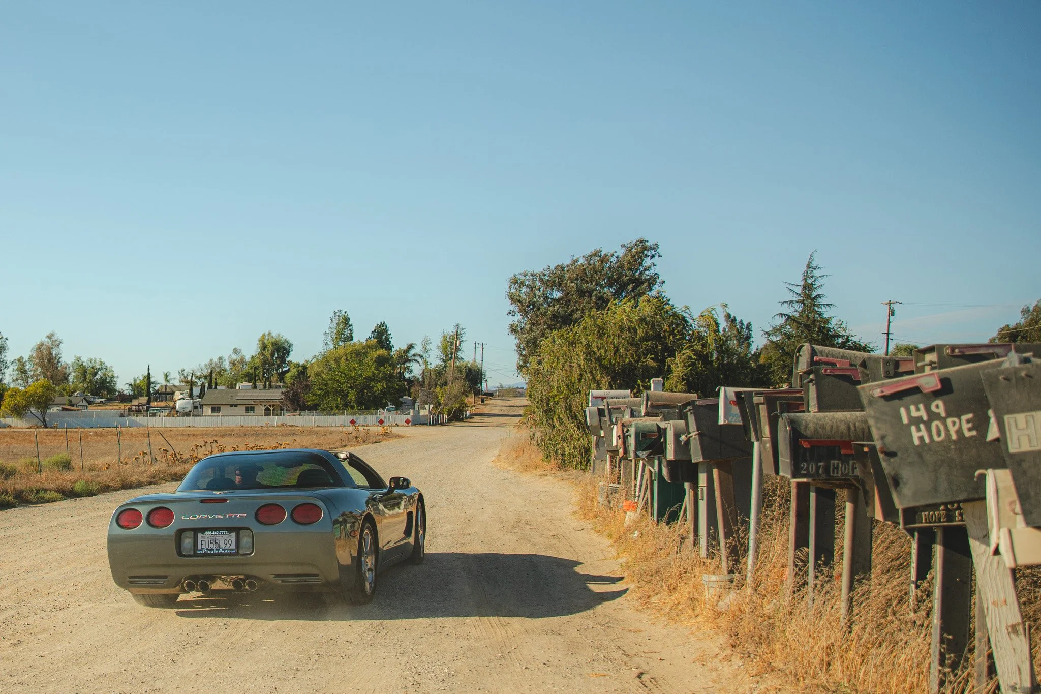 A silver Chevrolet Corvette parked on a dirt road with a row of mailboxes to the right and trees, houses, and clear blue sky in the background.