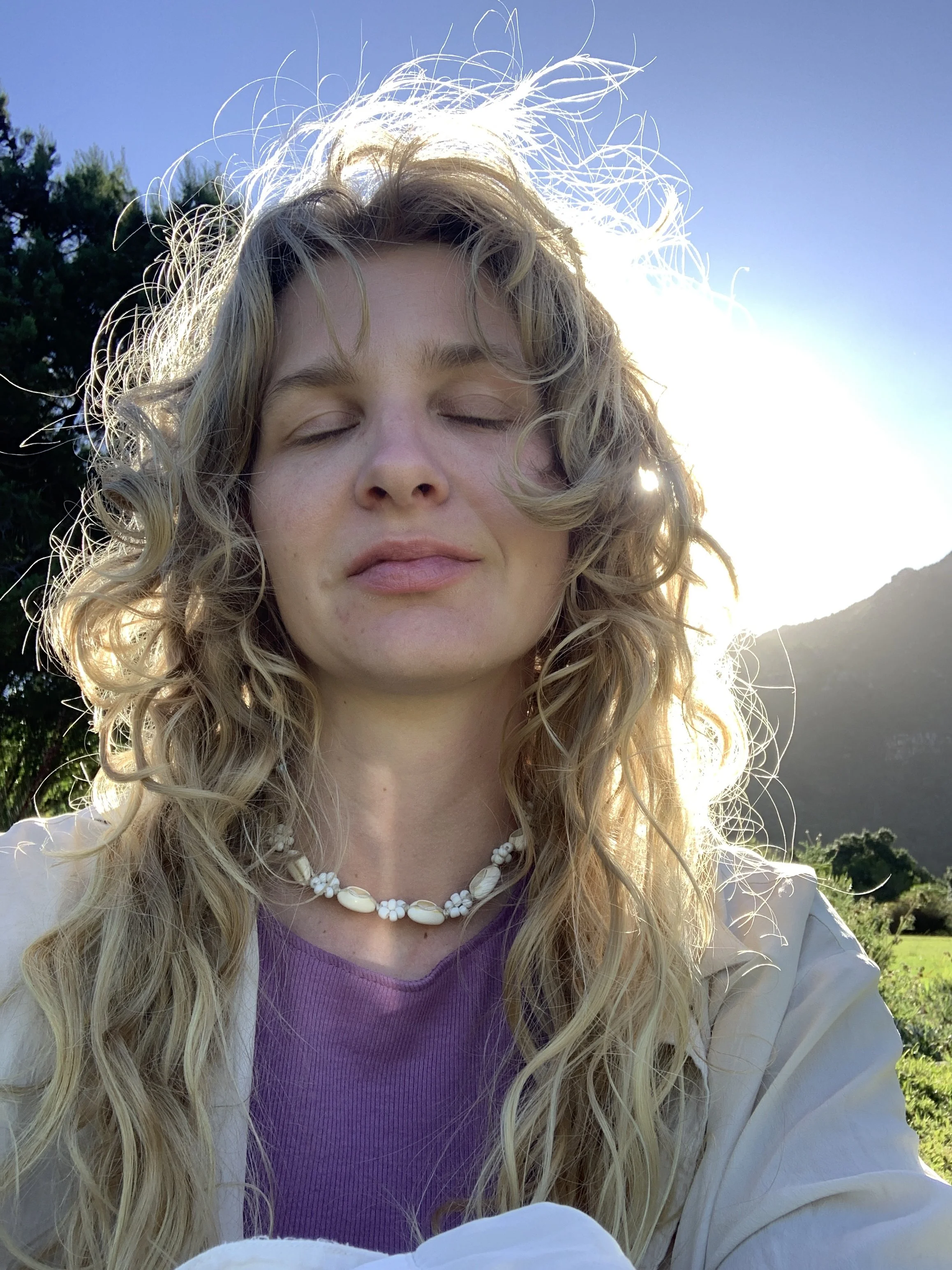 Junge Frau mit geschlossenen Augen, lange blonde, lockige Haare, trägt eine Perlenkette, blauer Himmel, Sonne im Hintergrund, Natur im Freien.
