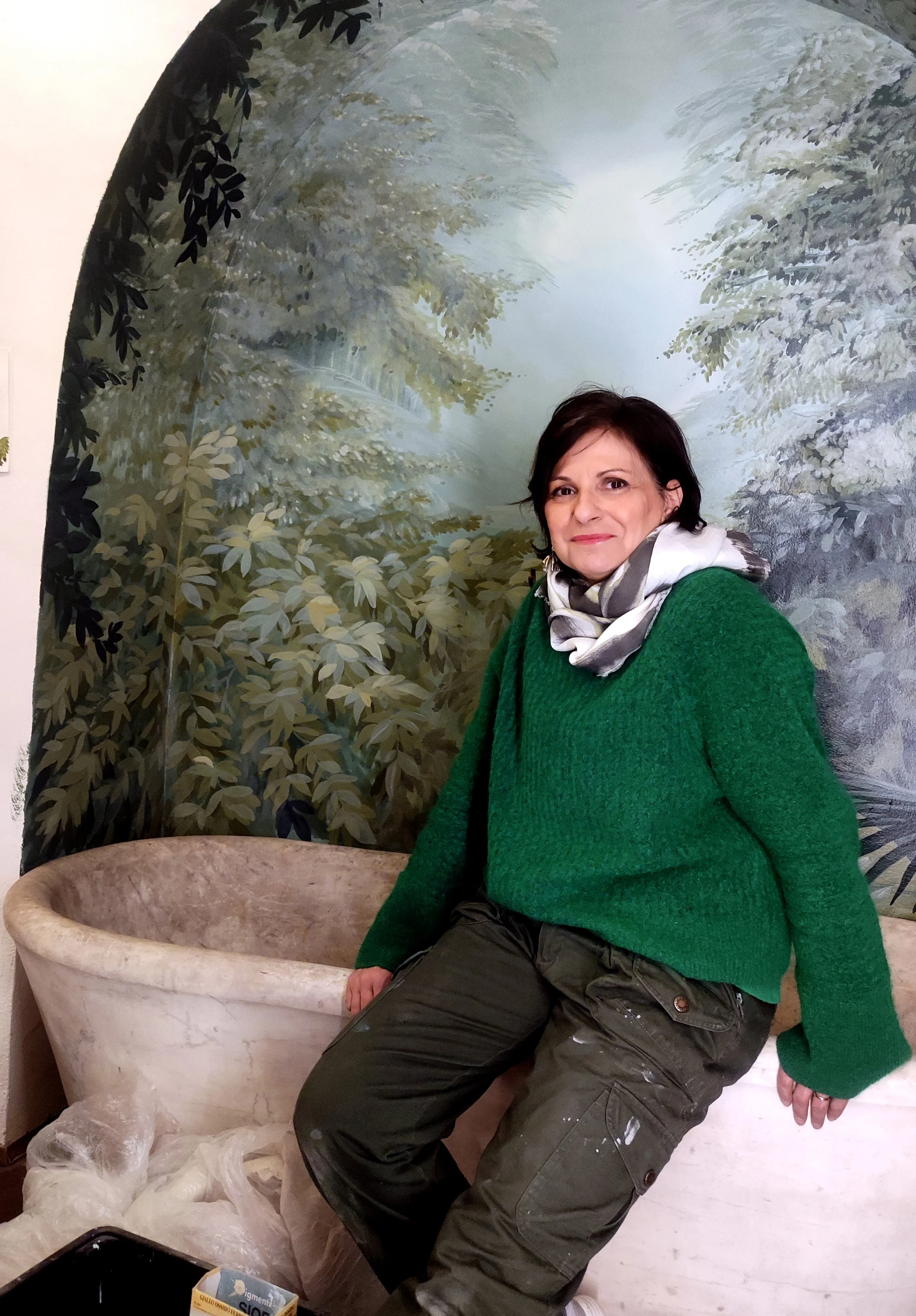 Laura Piacquadio seduta sul bordo di una grande vasca in pietra, davanti a un murale raffigurante alberi e vegetazione.