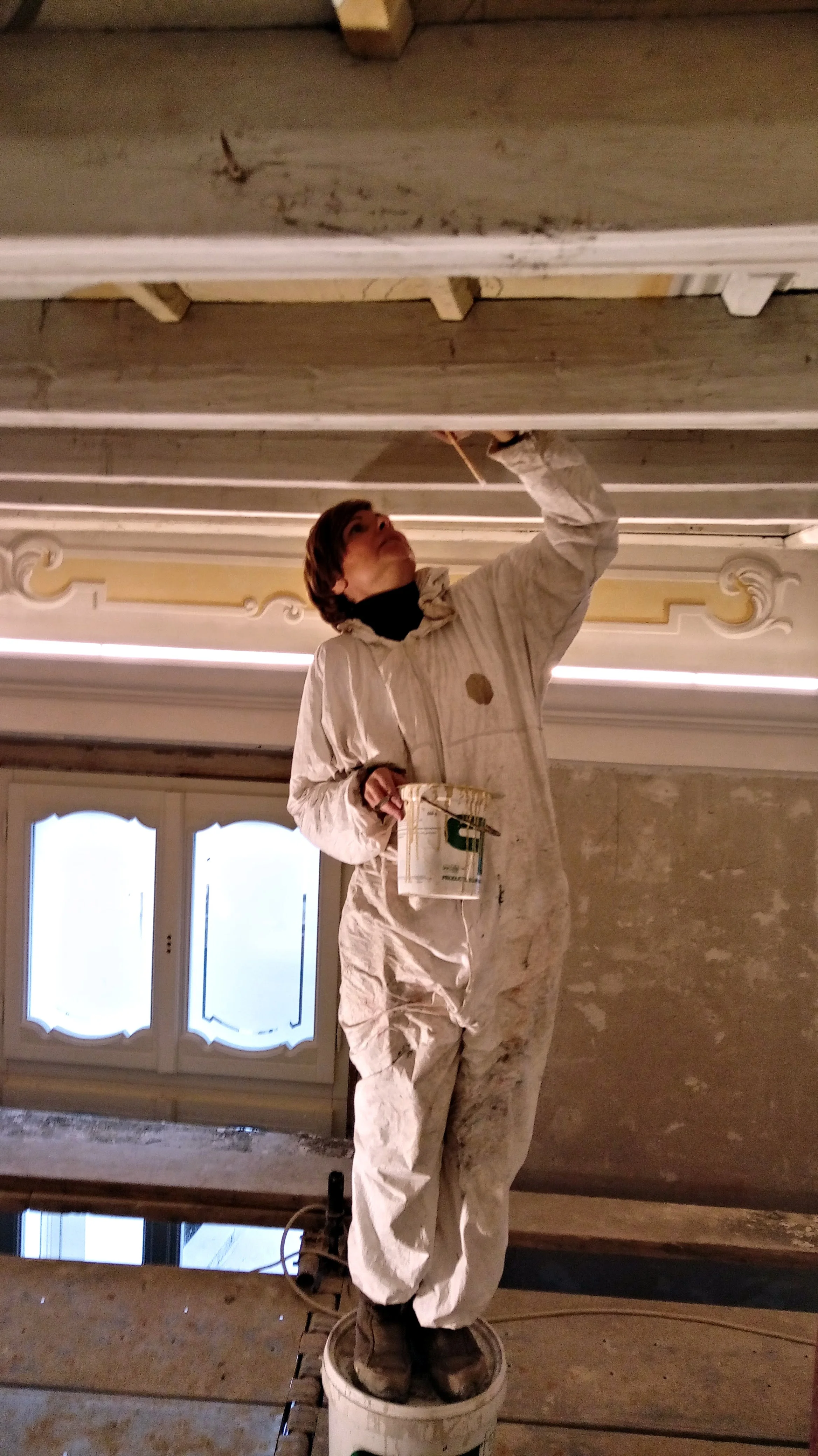 Donna al lavoro in un ambiente in ristrutturazione, mentre esegue un intervento pittorico sul soffitto.