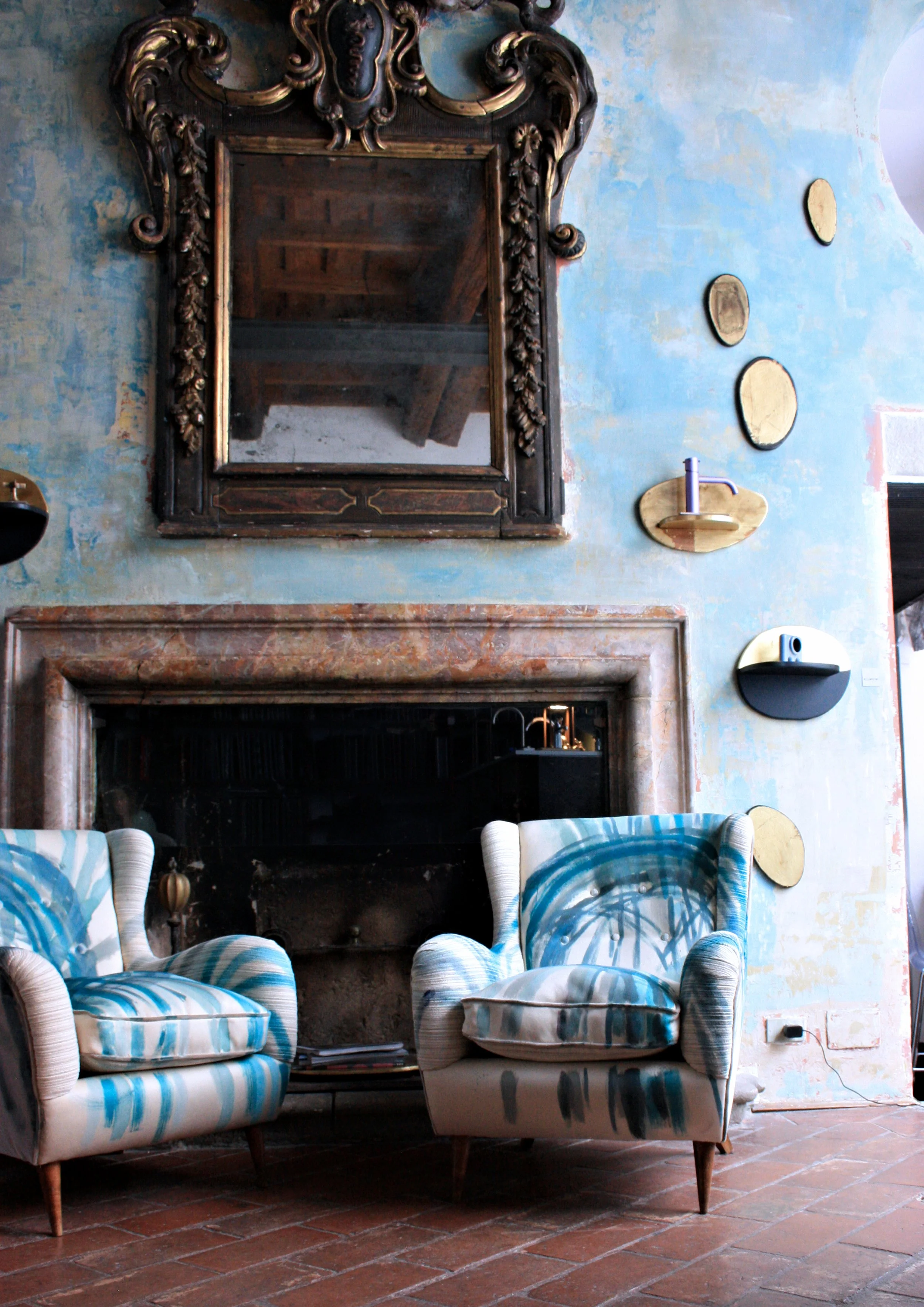 Soggiorno con grande specchio decorativo sopra il camino, parete blu con finitura materica, specchi decorativi e poltrone rivestite con tessuti decorati.