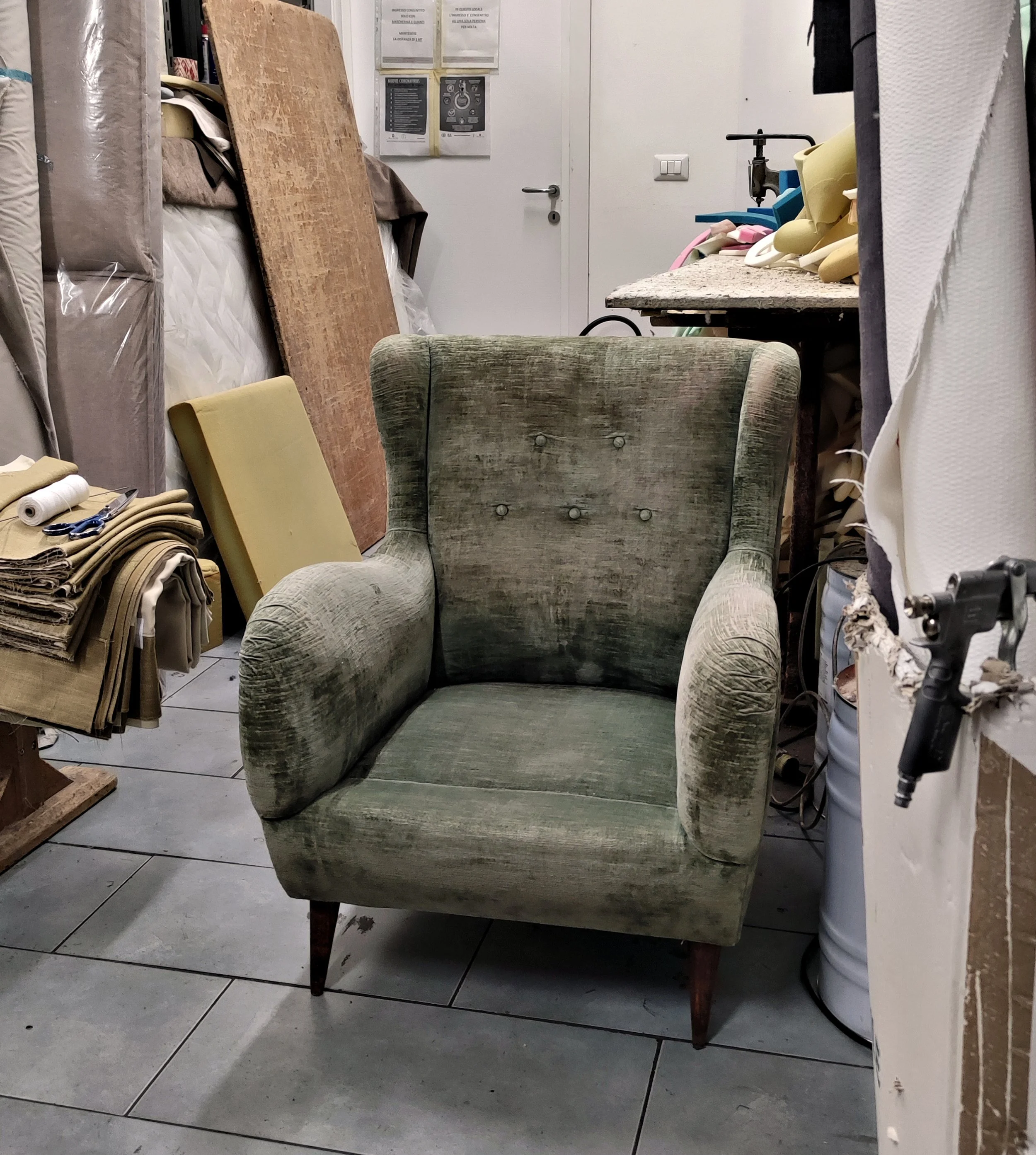 A vintage green upholstered armchair with wooden legs in a cluttered room surrounded by fabric rolls, a wooden board, and sewing supplies. A white door with information posters is visible in the background.