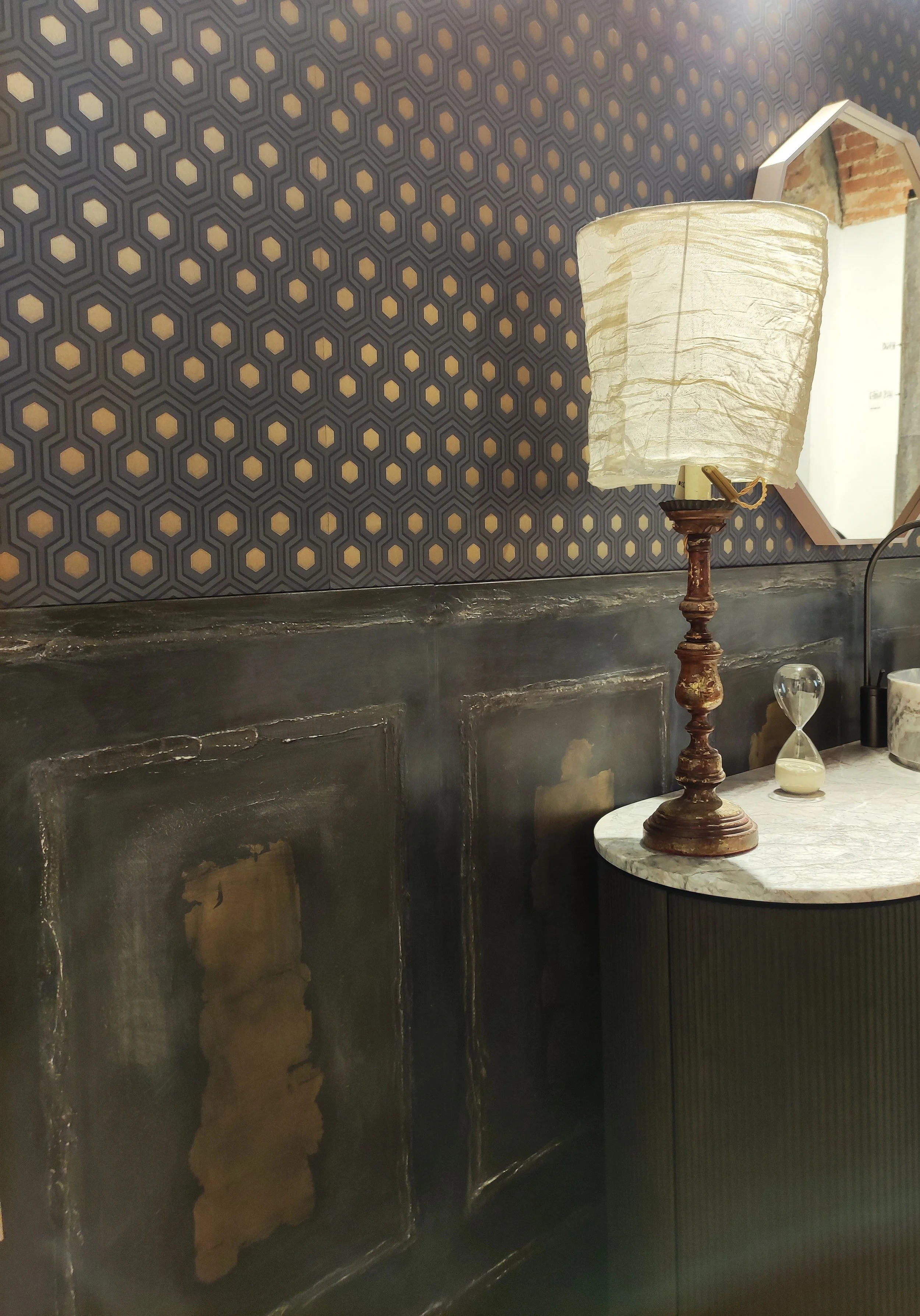 Tavolino con piano in marmo, lampada vintage e clessidra, davanti a una parete decorata con motivo esagonale.