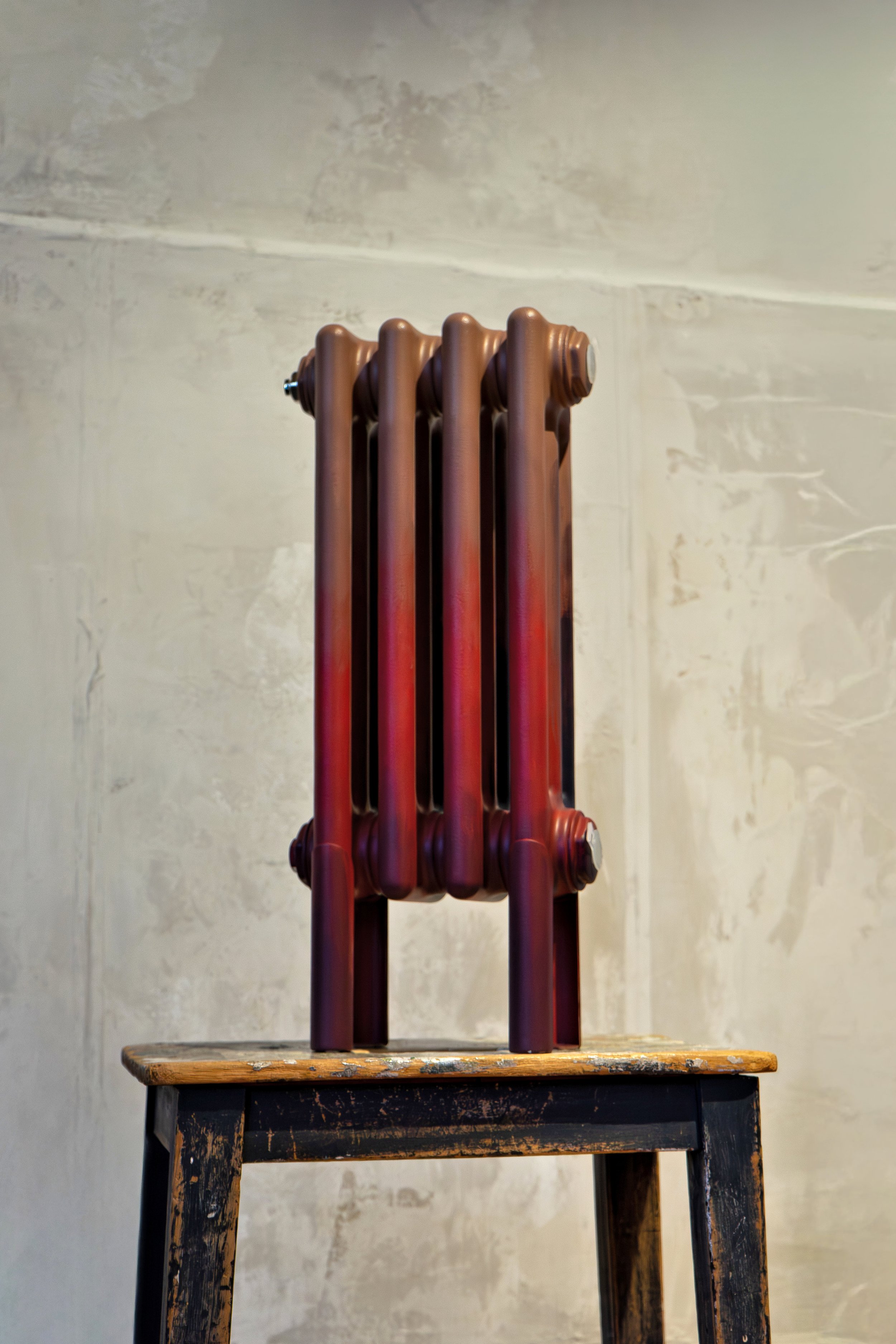 Radiatore vintage in ghisa dipinto nei toni del rosso-bruno, appoggiato su un vecchio sgabello in legno.