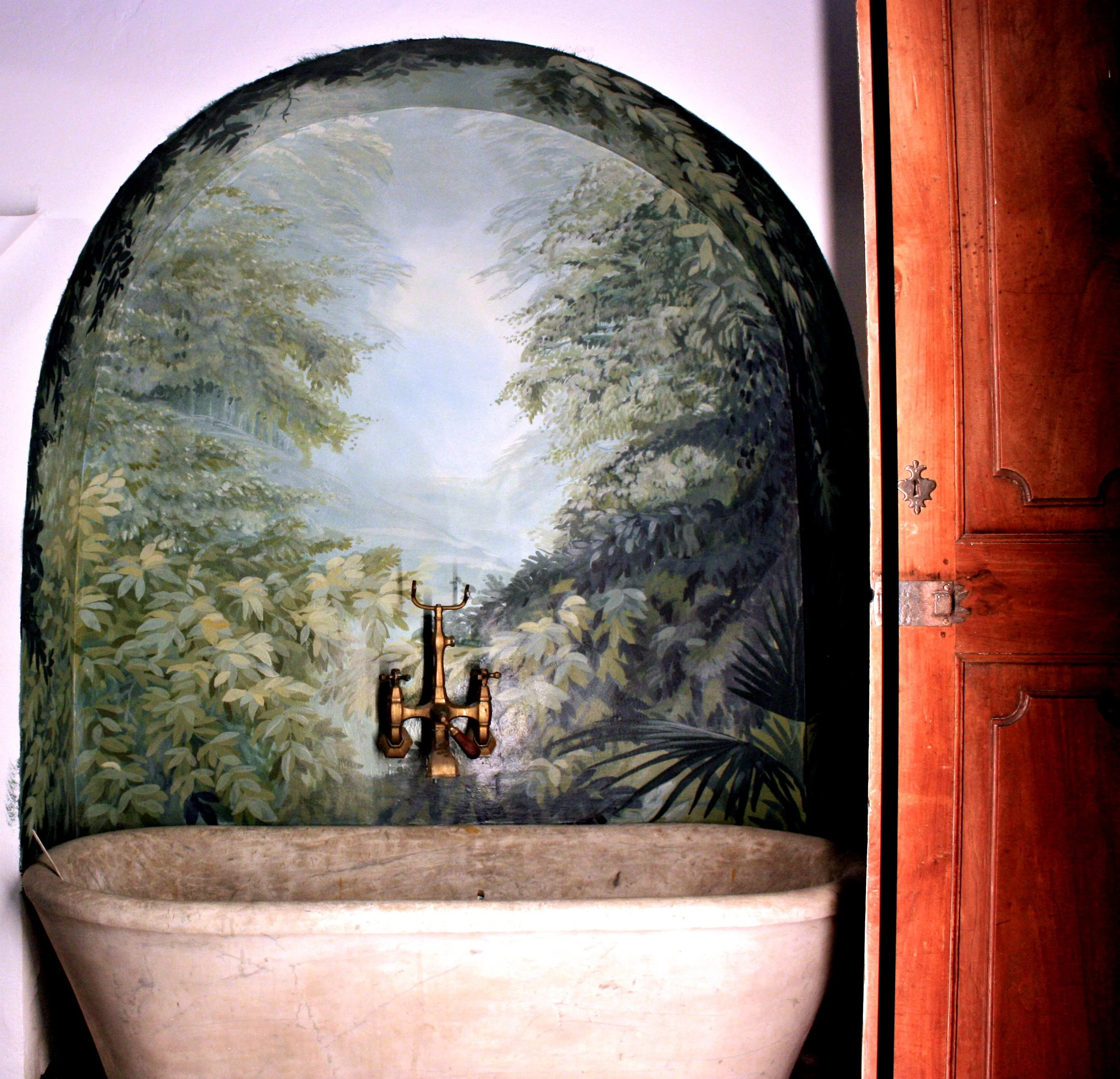 Vasca vintage in pietra con rubinetteria in ottone davanti a un paesaggio boschivo dipinto.