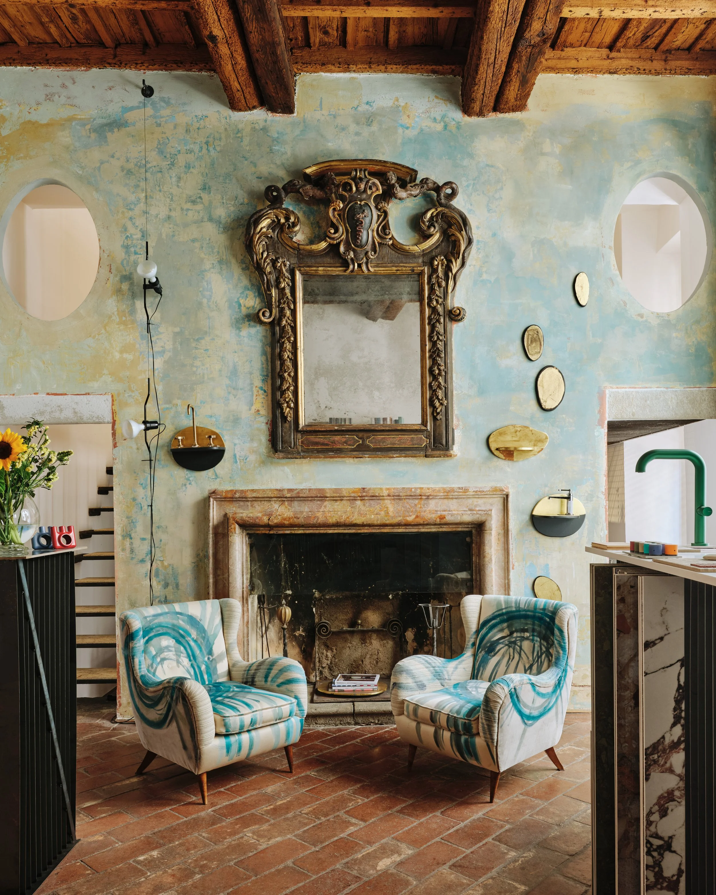 Living room with two patterned armchairs, a fireplace with a mirror above it, and rustic wooden ceiling beams. The room has textured, worn walls with distortions and circular decorations, and adjacent rooms are visible through arches.