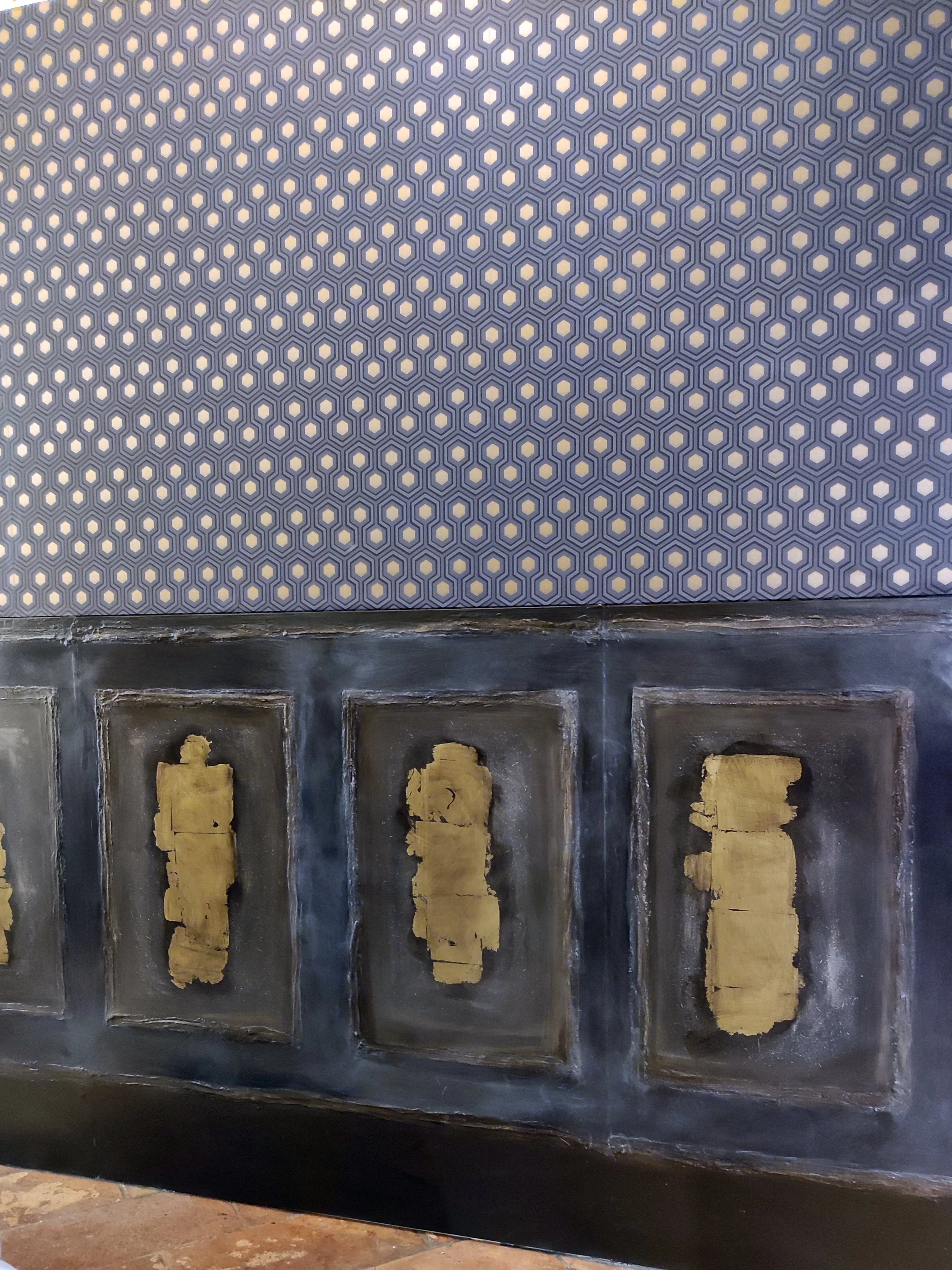 Parte di una parete decorata con un motivo esagonale nella parte superiore e un pannello in legno con dettagli dorati nella parte inferiore.