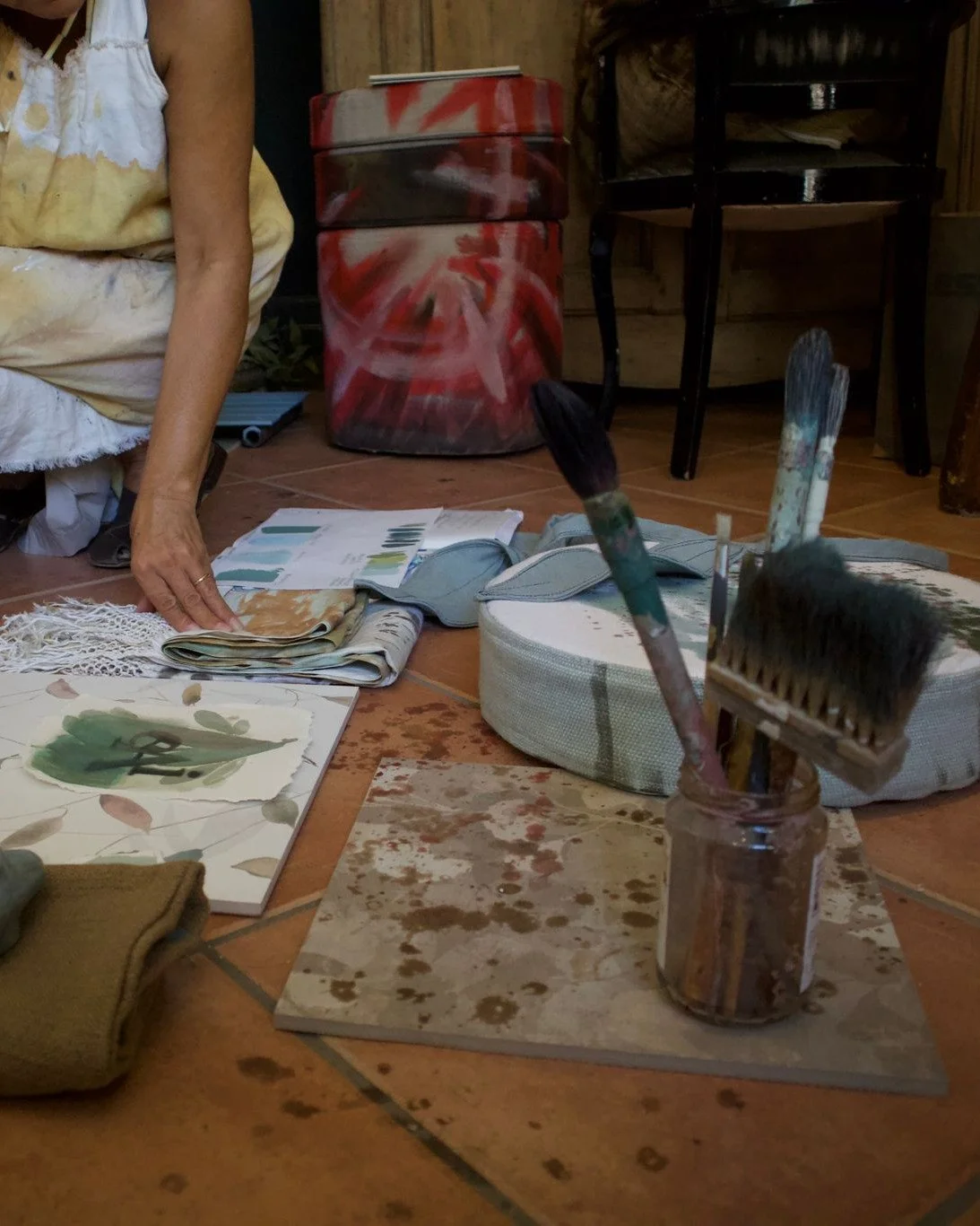 Atelier dell’artista con colori, pennelli, carta e tessuti sul pavimento. Una persona è inginocchiata mentre sistema i materiali.