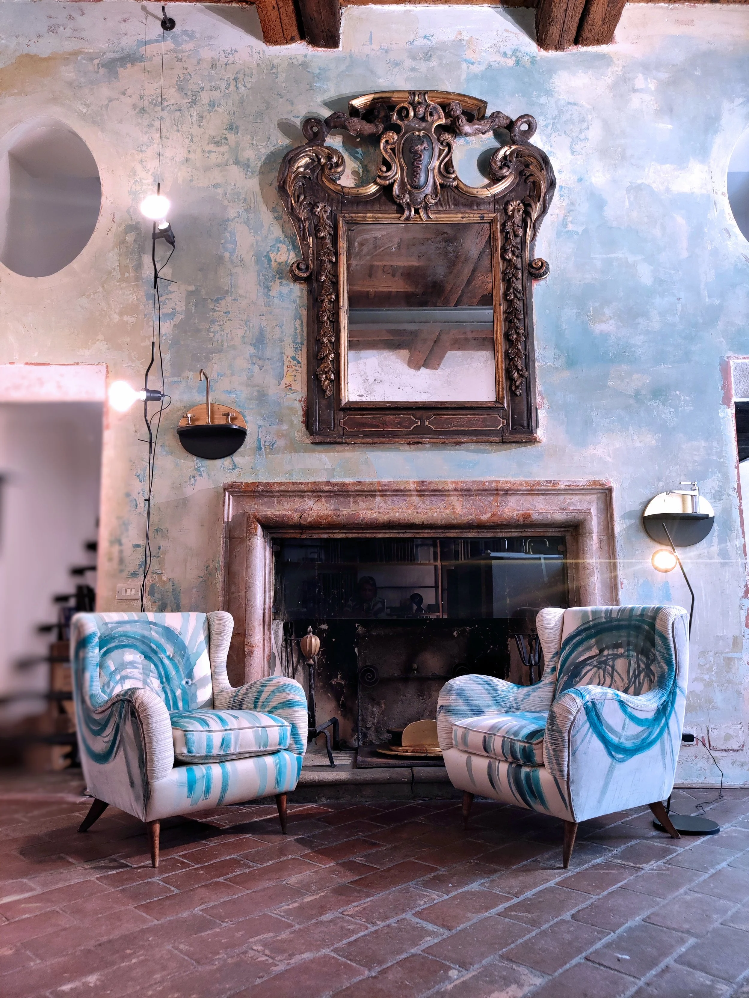 Due poltrone vintage rivestite con motivo blu e bianco davanti a un camino in mattoni e a un grande specchio decorativo.