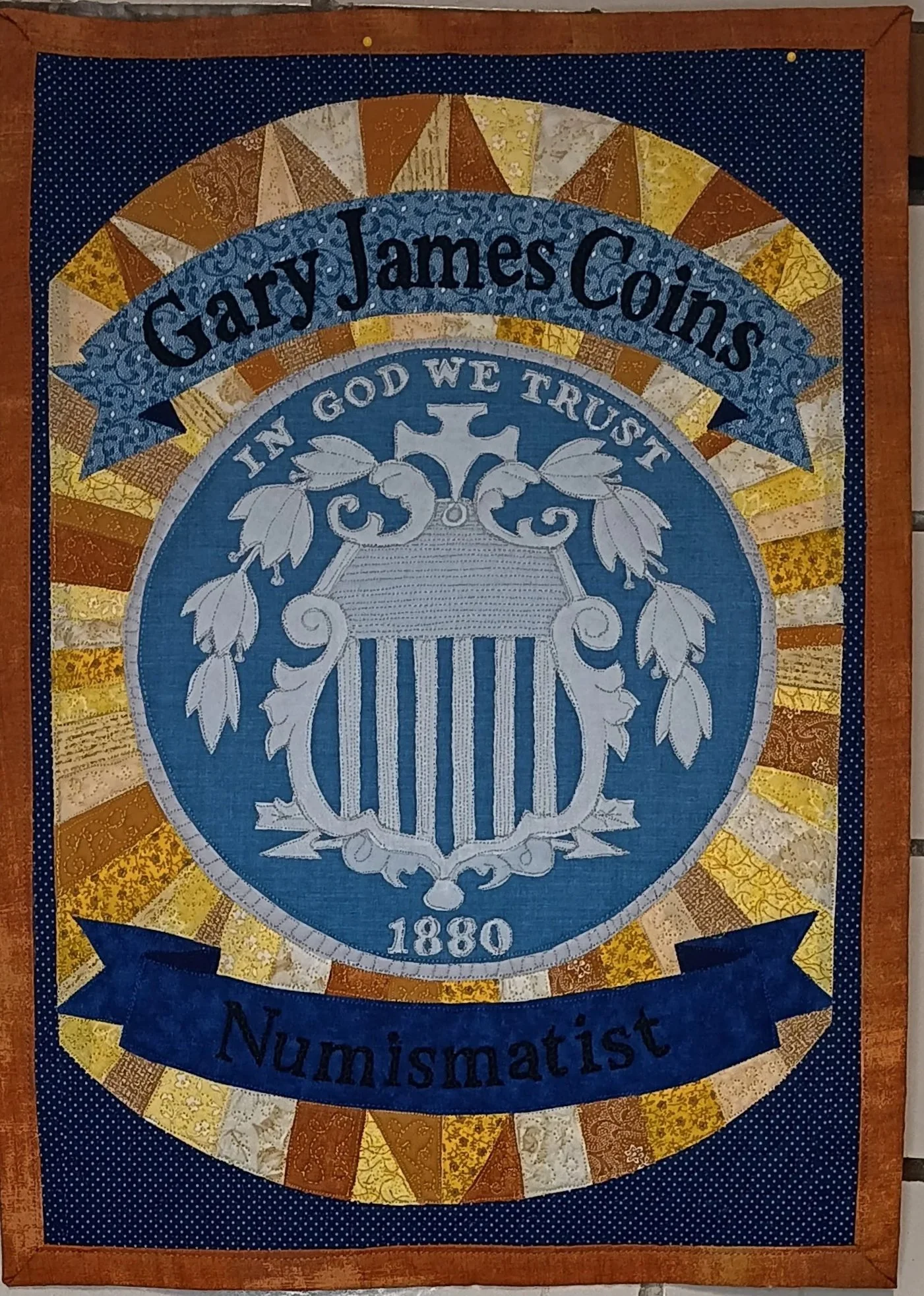 A quilted banner displaying the name Gary James Coins, with the words 'In God We Trust' and '1880',  a shield nickel surrounded by a decorative patchwork border in shades of brown, yellow, and blue.
