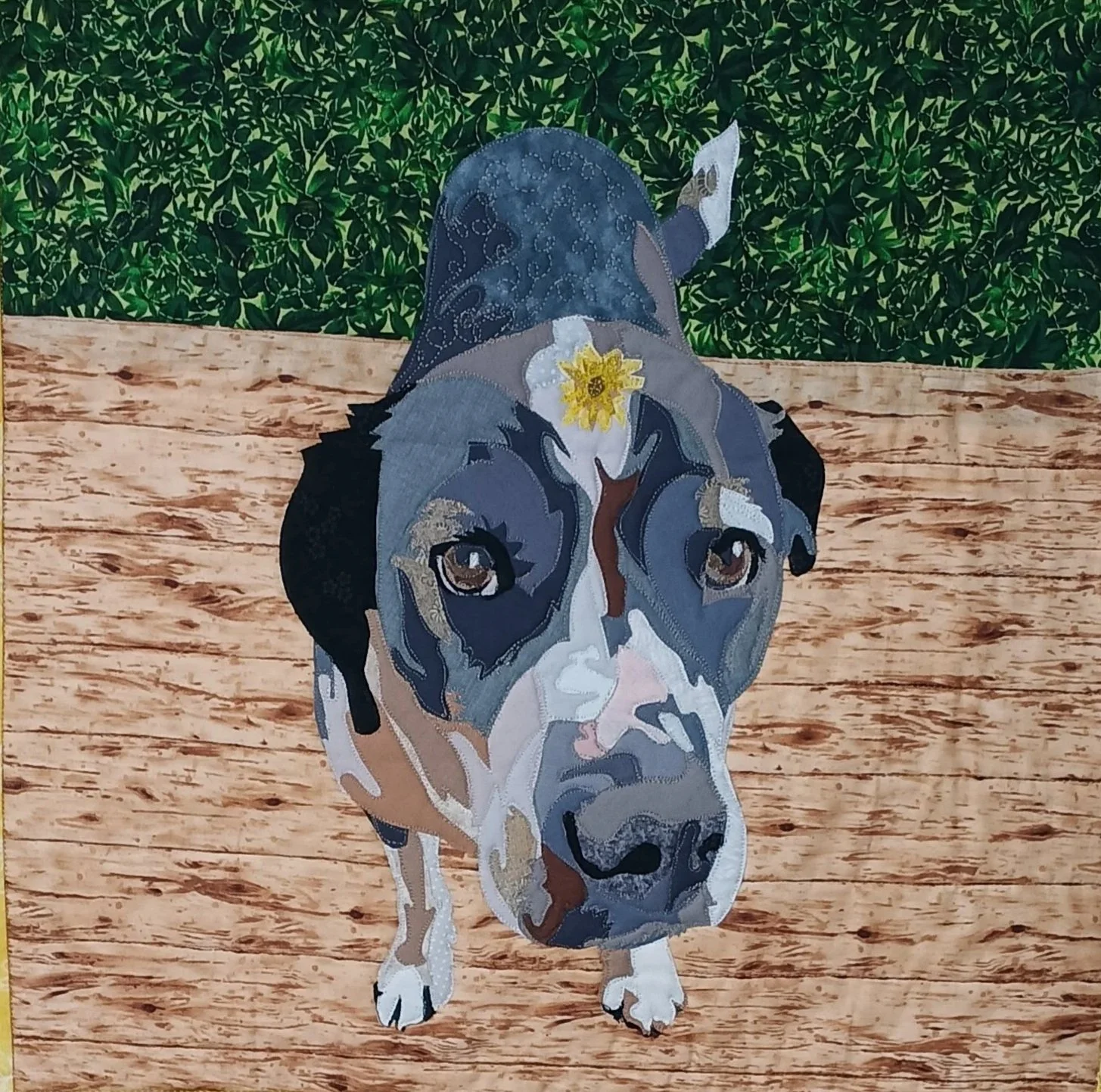 A detailed fabric artwork of a dog's face with a dandelion on her head, with a background of green bushes and a wooden surface.