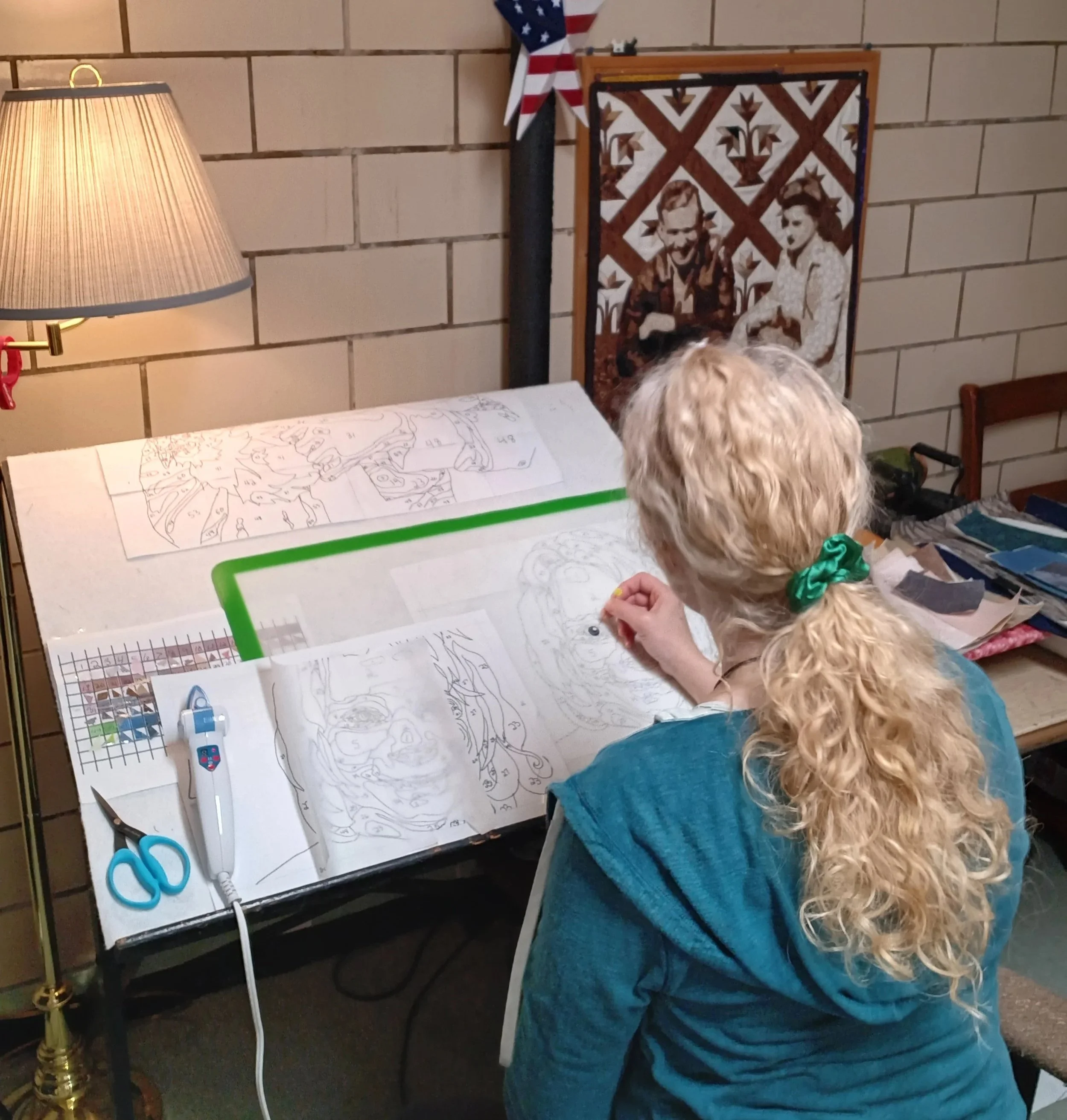 A person with long curly blonde hair sits at a table, working on a  The table has various tools and supplies, including white and blue scissors, a glue gun, and some paper with sketches or outlines. Behind them, there is a quilted wall hanging and a lit table lamp with a beige shade. The setting appears to be indoors, possibly a craft room or workshop.
