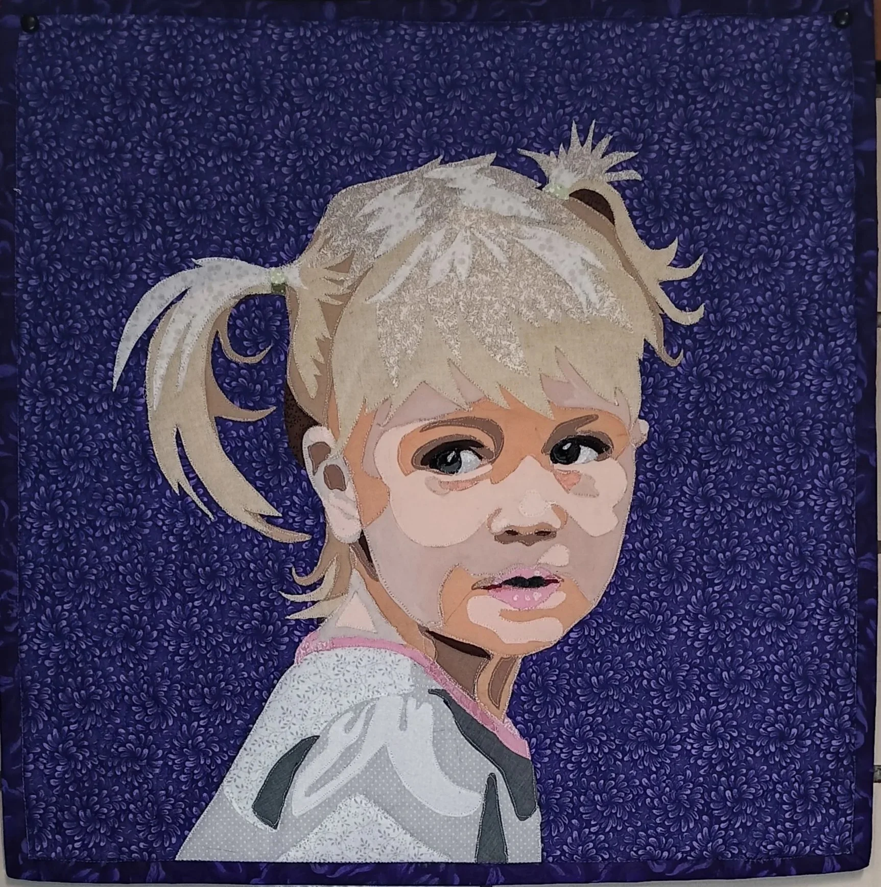 A close-up of a quilting artwork featuring a portrait of a young girl with blonde hair, large expressive eyes, and a serious expression, set against a dark blue, patterned background.