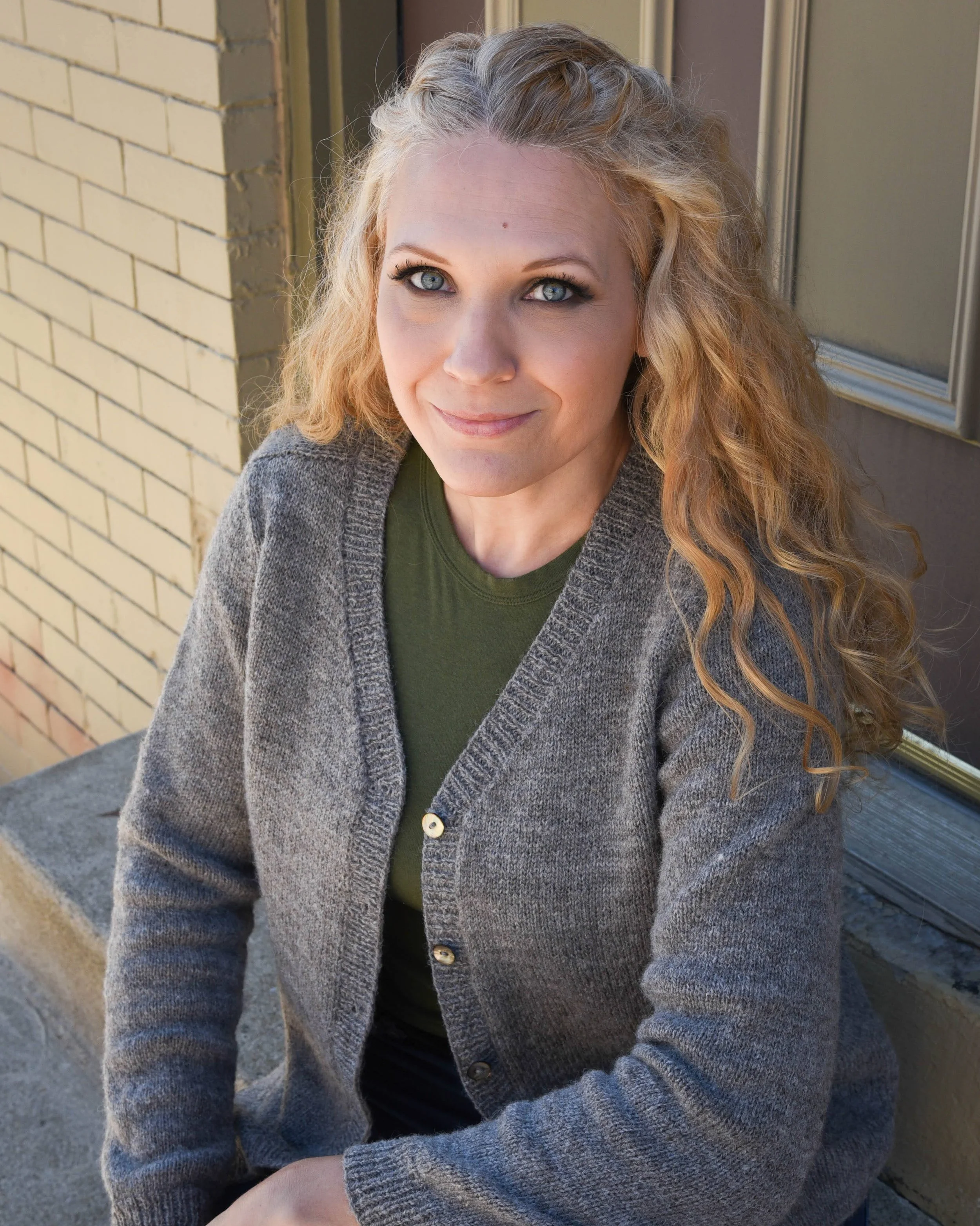 A woman with curly blonde hair and blue eyes smiling outdoors, wearing a green top and gray cardigan, seated beside a brick wall and a window.