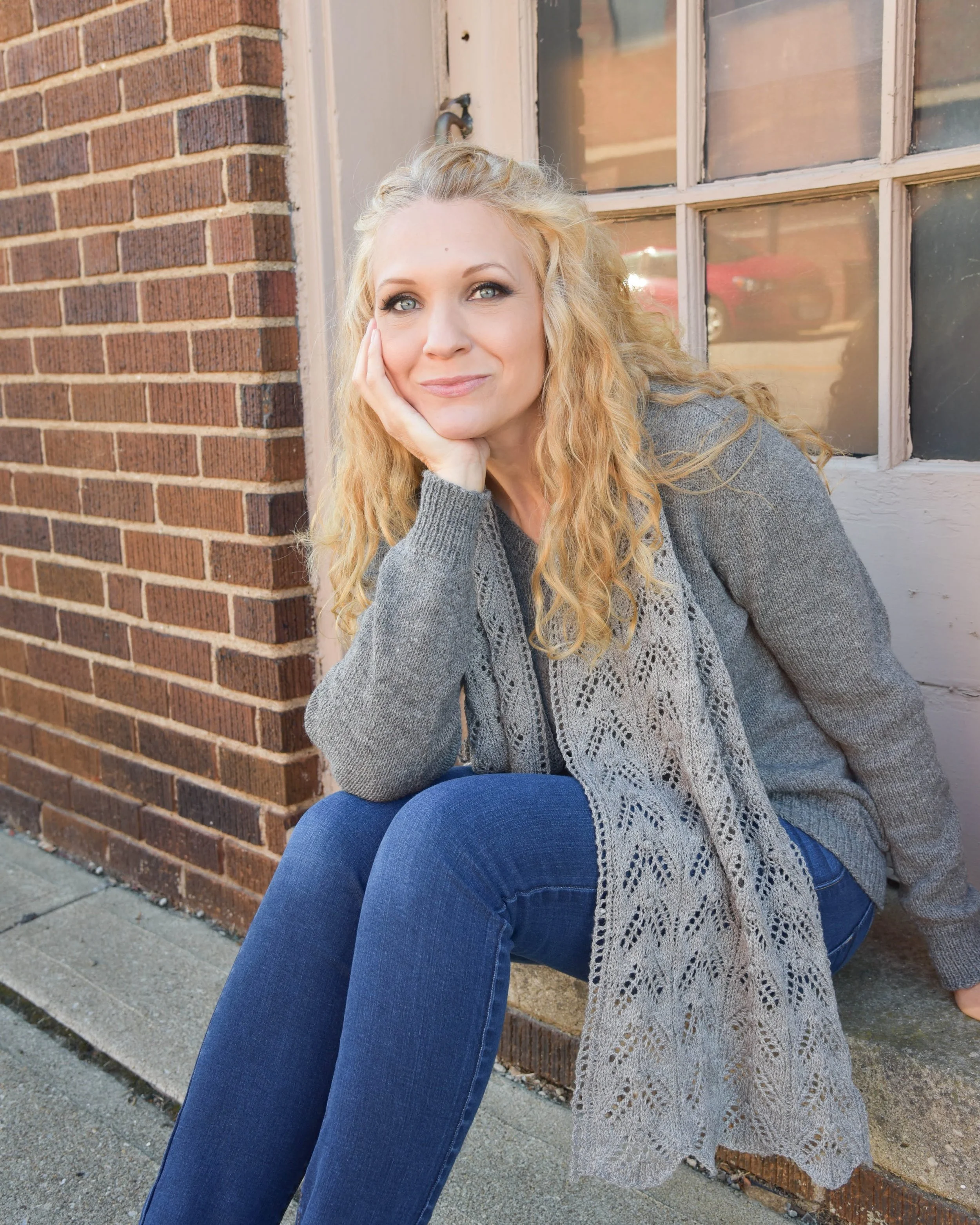 A woman with curly blonde hair sitting outdoors against a brick wall with a window, wearing a gray sweater and blue jeans, resting her chin on her hand and smiling softly.