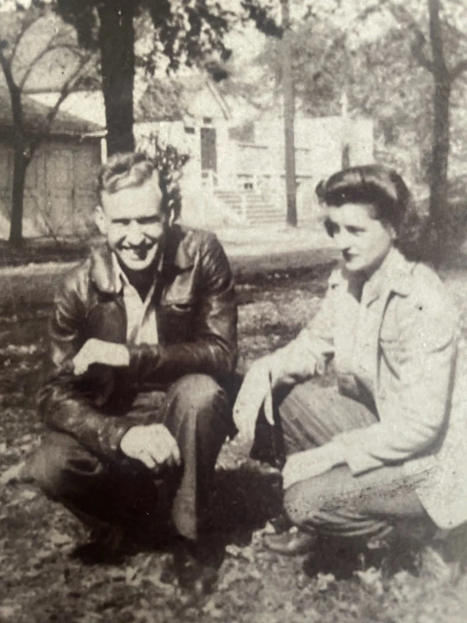A young couple sitting outdoors on grass, smiling and posing for the camera, with trees and a building in the background.
