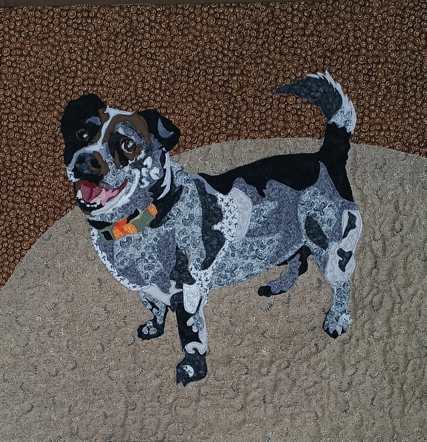 A quilted fabric artwork of a speckled dog, lying on a patterned surface with a brown patterned background.