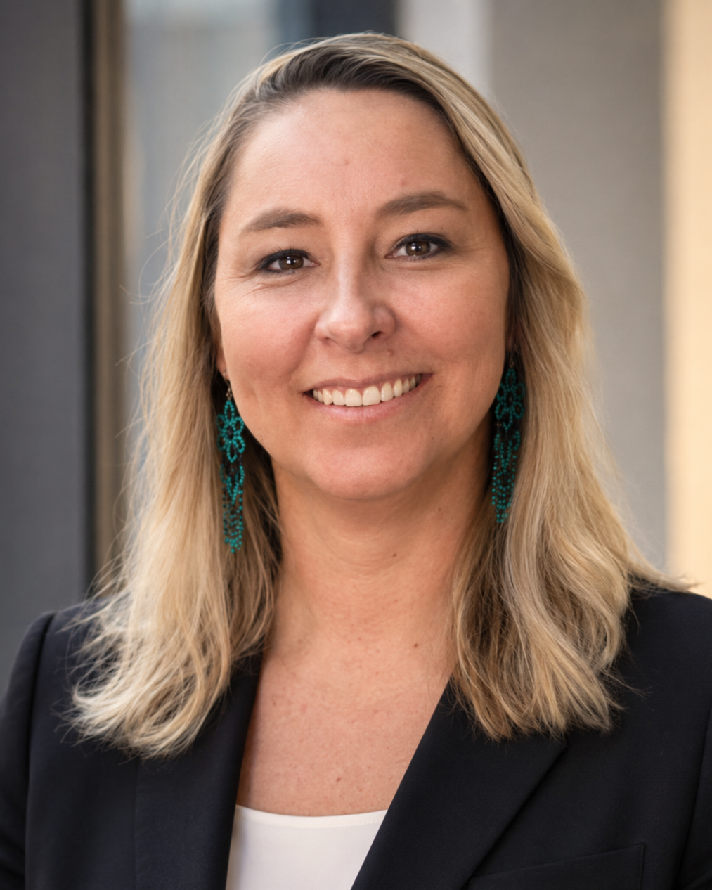 A smiling woman with blonde hair wearing teal earrings and a black blazer.