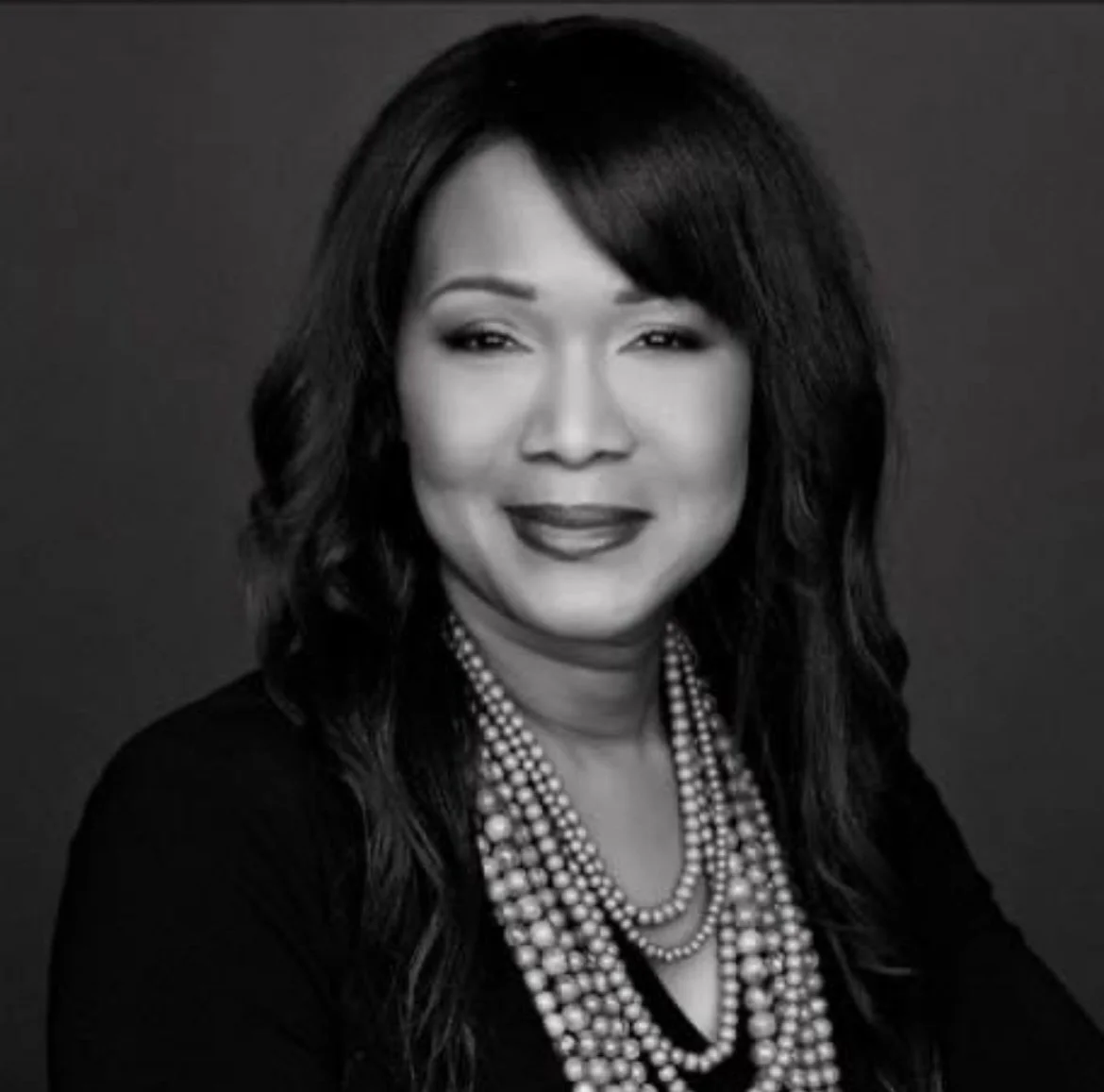 Black and white photograph of a woman with wavy hair wearing a dark top and a multi-strand pearl necklace, smiling at the camera.