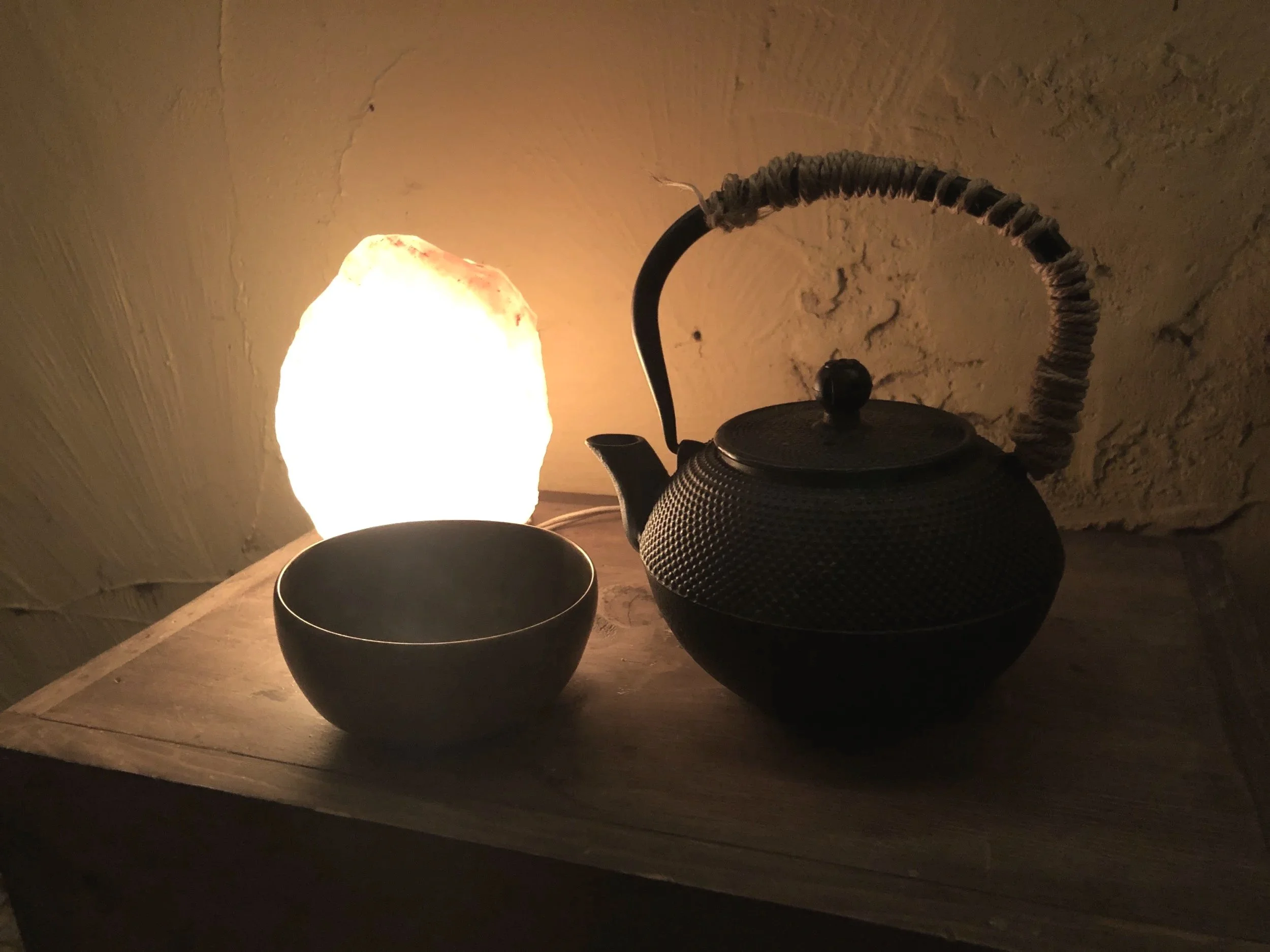 A black teapot with a textured surface, a black kettle, and a black bowl or cup on a wooden surface against a textured wall. A lamp with a warm light is partially visible on the left.