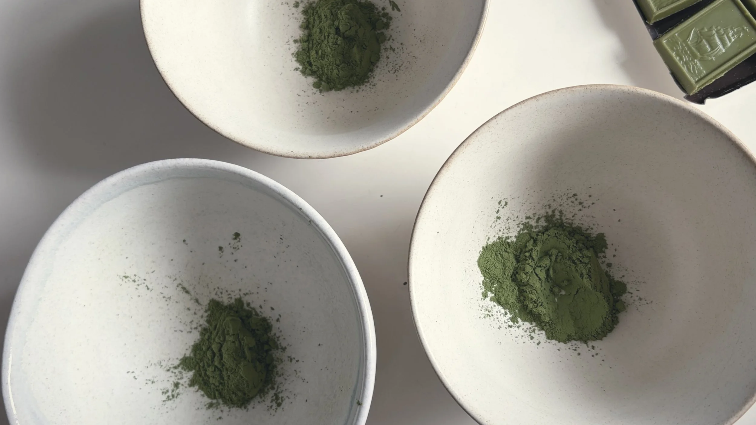 Small white bowls containing green matcha powder, with a green matcha tea stamp visible on one of the bowls.