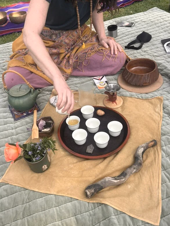 Person sitting on a blanket with a tea set, flowerpot, and decorative items for a tea ceremony outdoors.