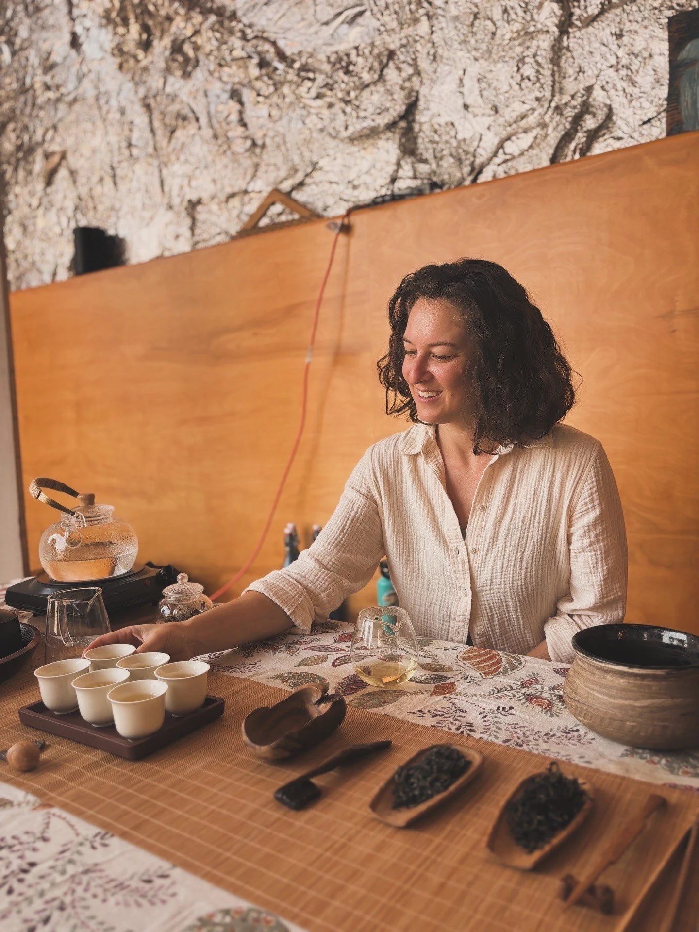 Woman seated at a table with tea cups, teapot, and various traditional objects, engaging in a tea ceremony in a cozy room with wooden decor and a decorative table runner.