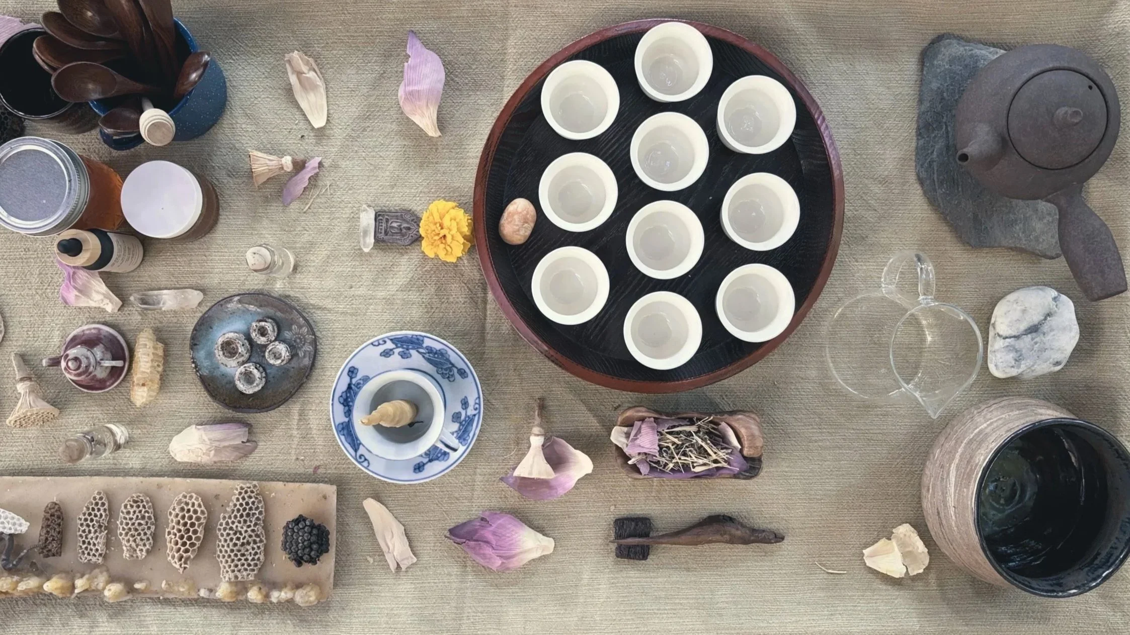 A table with various objects including small white cups, dried flower petals, rocks, a teapot, and decorative items like honeycomb-shaped pieces, dried herbs, and small bottles.